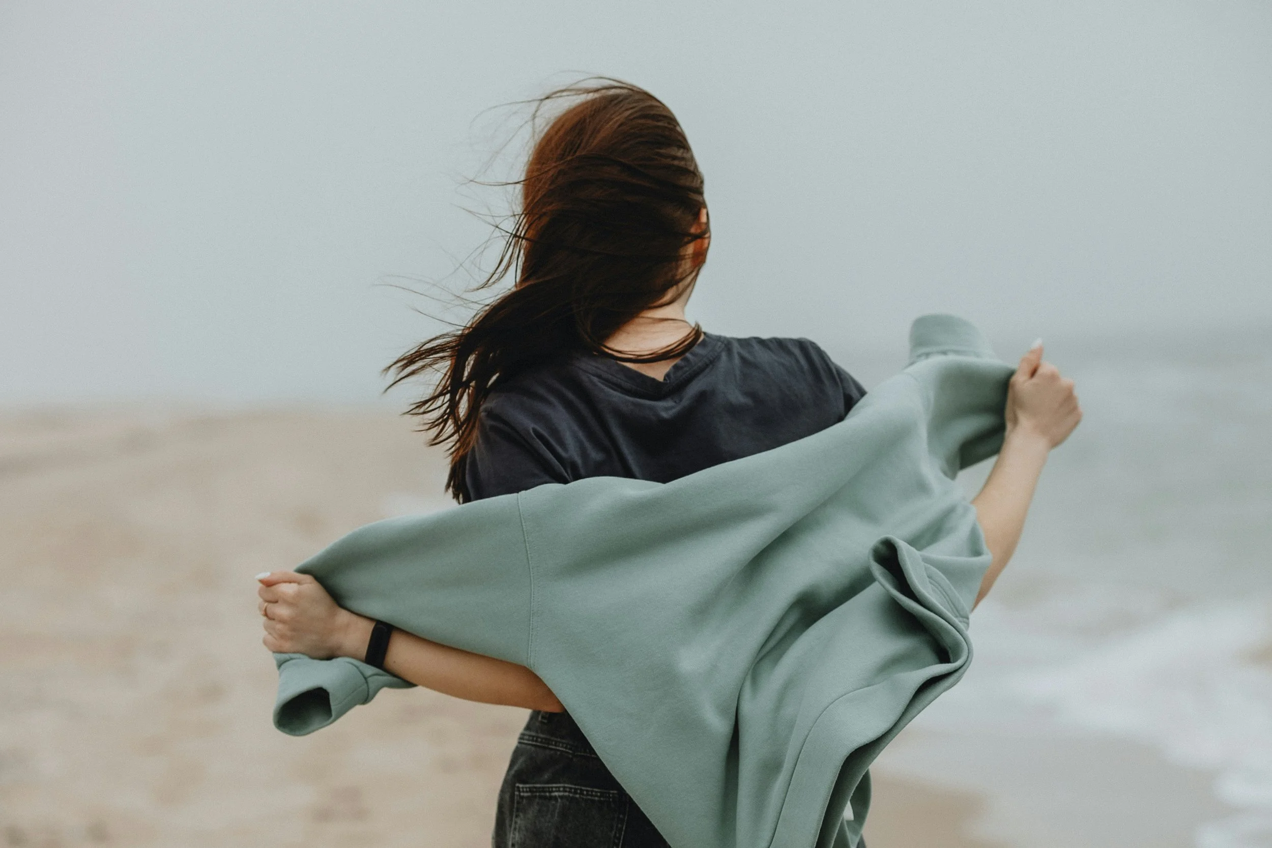 A woman standing on the beach, holding a light green jacket, with wind blowing her brown hair back, overlooking the ocean.