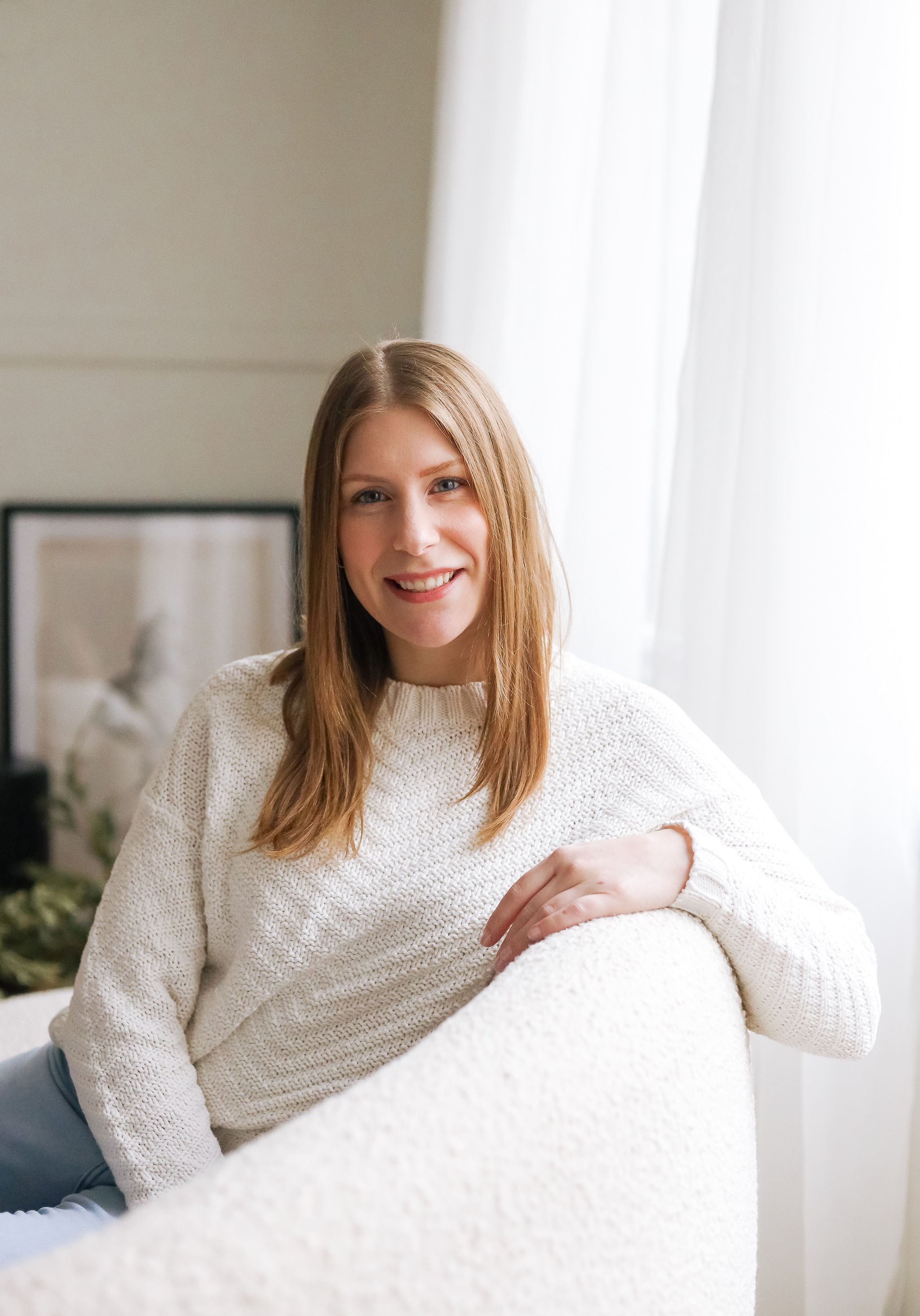 Smiling woman with long brown hair, wearing a white knit sweater, sitting on a white sofa in a bright room with natural light, white curtains, and a framed artwork in the background.