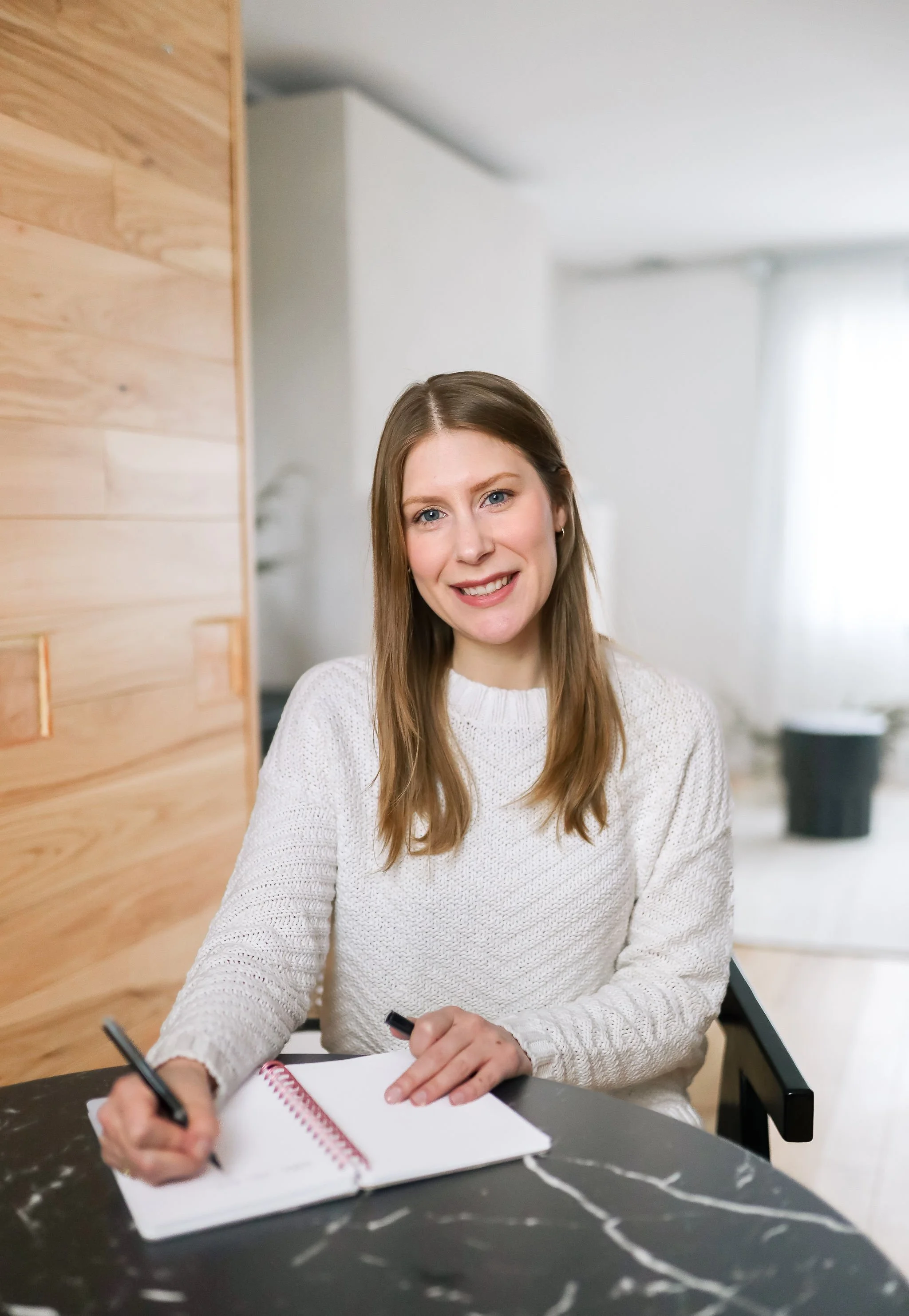 A young woman with long blonde hair, wearing a white sweater, sitting at a black marble table, smiling, holding a pen over an open notebook.
