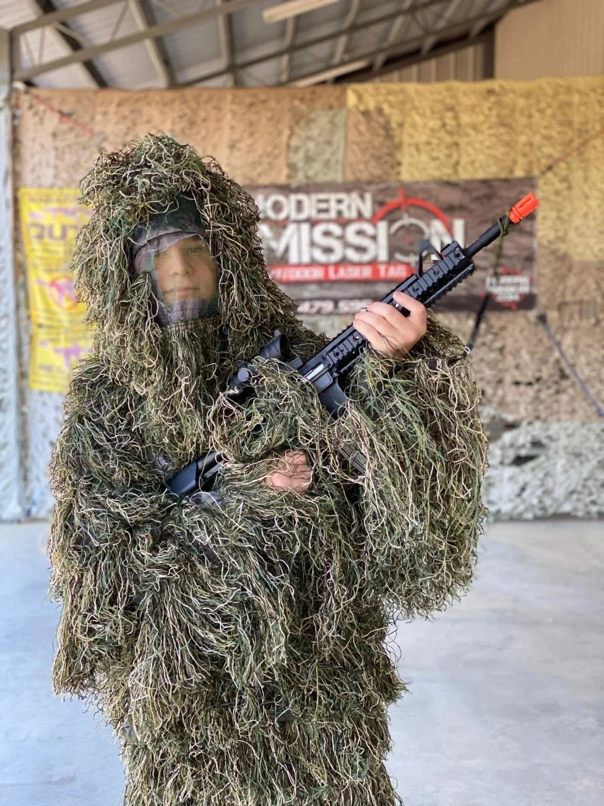 A person dressed in a camouflage ghillie suit holding a toy or replica rifle at an indoor laser tag center.