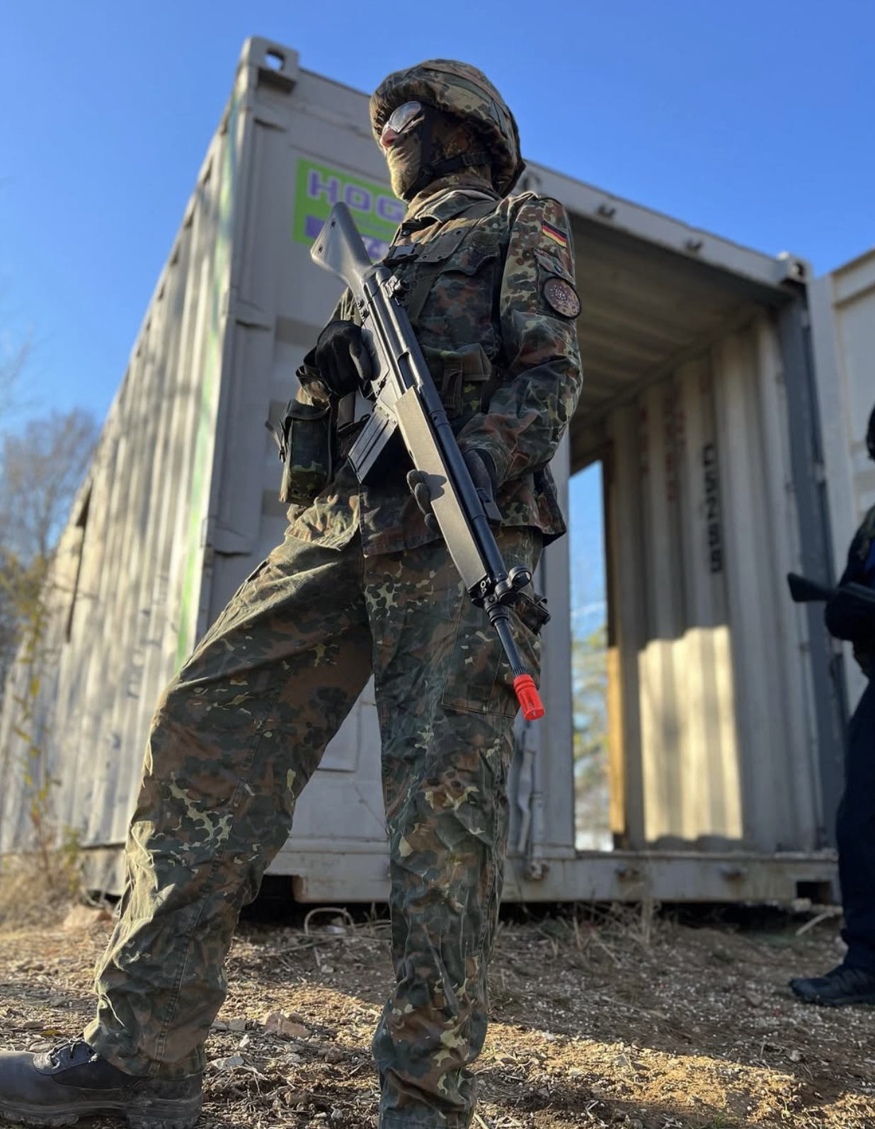 A person dressed in camouflage military gear, holding a rifle, standing outdoors in front of a shipping container, with a clear blue sky in the background.