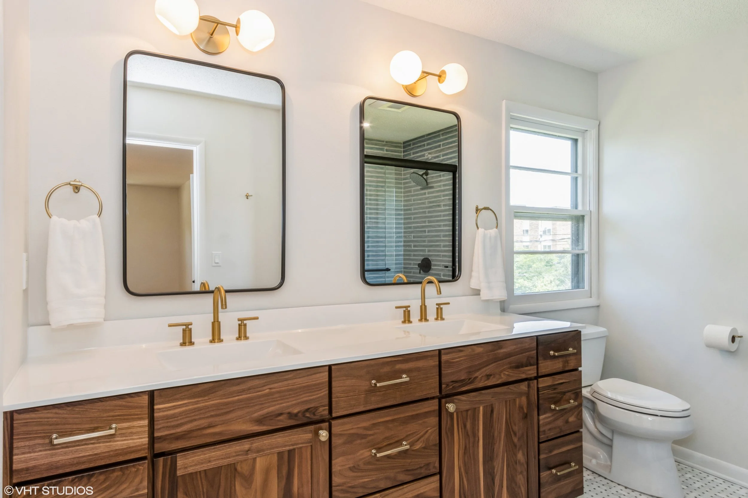 Bathroom with double vanity, gold fixtures, two large mirrors, and a window on the right. The vanity has wooden cabinets and a white countertop. A towel hangs next to each mirror, and in the reflection, a shower with gray tiles and a glass door is visible.