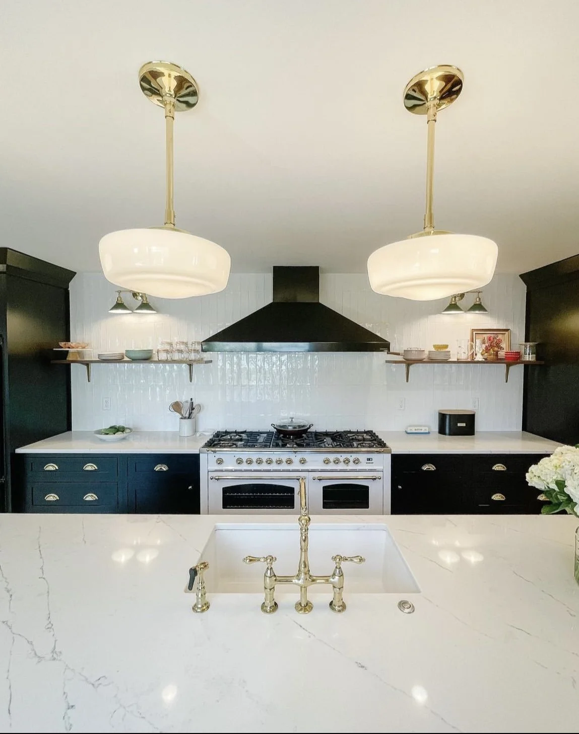 Modern kitchen with white marble island, black cabinets, and a stainless steel stove. Two large white pendant lights hang from the ceiling over the island, with a brass faucet and sink. Shelves with dishes and decorative items are on the wall behind the stove.