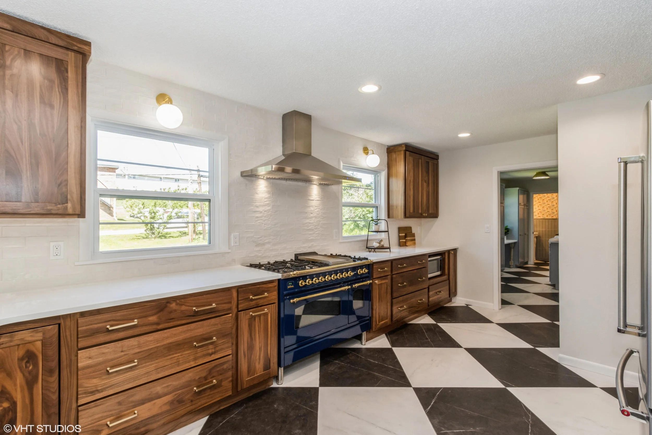 Kitchen with white walls, wooden cabinets, a blue stove, black and white checkered floor, windows, and a doorway to another room.
