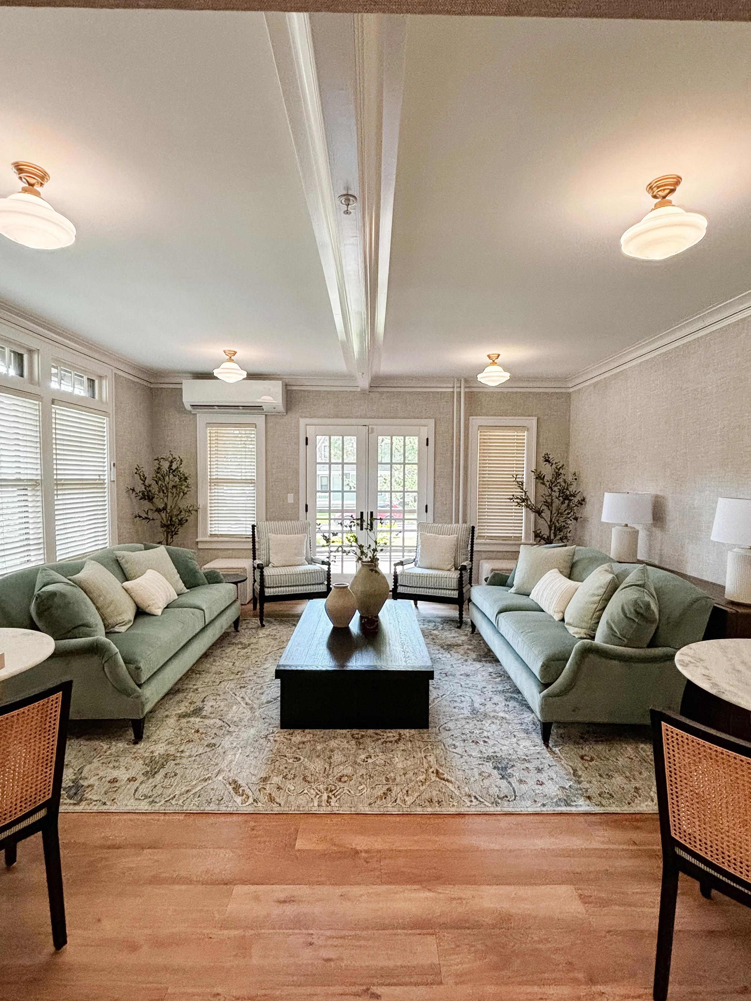 Living room with two green sofas, two armchairs, and a rectangular wooden coffee table with vases on top, beige walls, wooden flooring, large windows with white blinds, potted plants, and outdoor French doors.