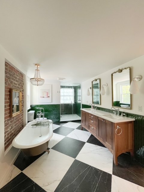 Bathroom with freestanding bathtub, double sink vanity, framed mirrors, brick wall accent, and checkered black and white tile floor.
