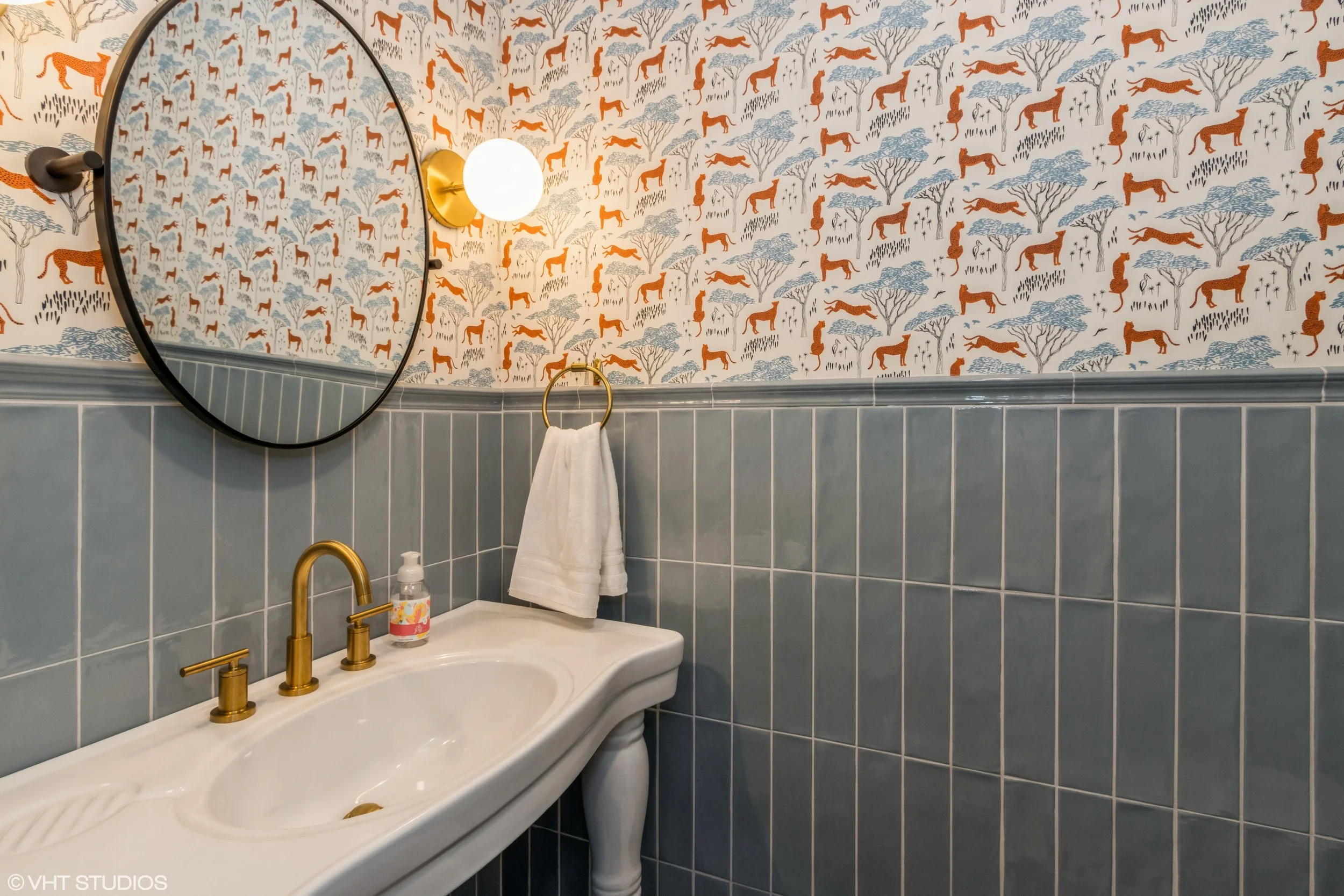 A bathroom corner with a blue tiled lower wall and a wallpapered upper wall featuring a pattern of orange and blue animals and trees. There is a white bathroom sink with gold fixtures, a round mirror, a wall-mounted light, a towel ring with a white towel, and a liquid soap dispenser on the sink.
