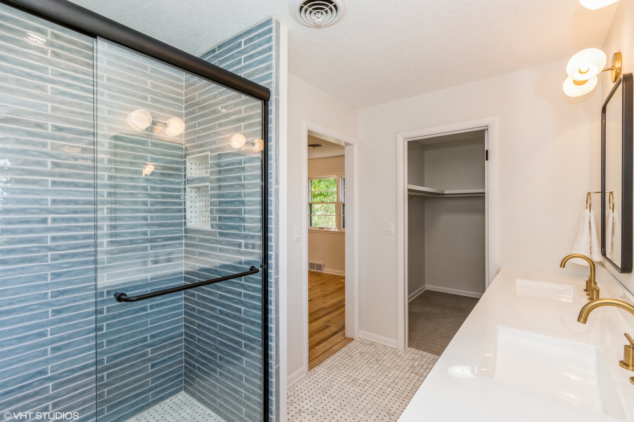Modern bathroom with a glass shower, white double sink vanity with gold fixtures, and an open closet with a shelf and hanging space.