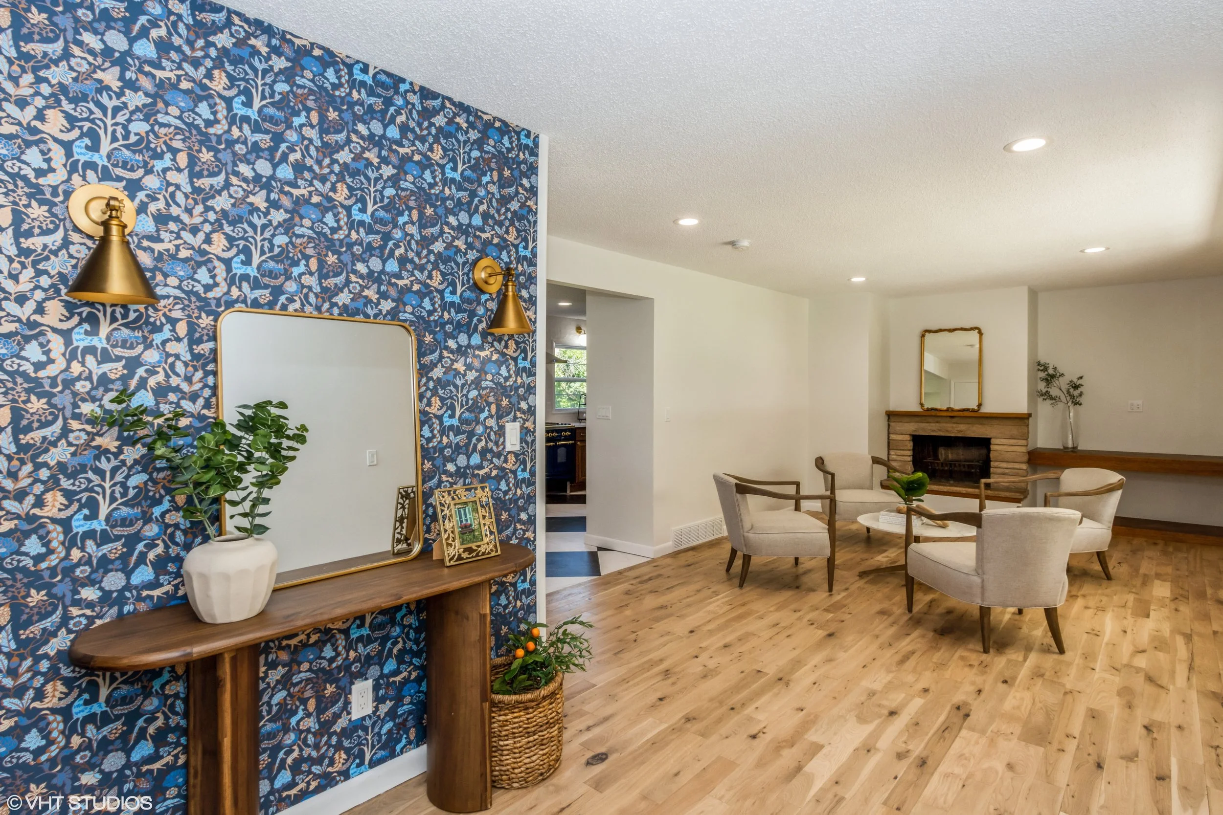 Living room with hardwood floors, white walls, a fireplace with a mirror above, four beige armchairs arranged around a small table with a plant, and a blue floral accent wall with two gold sconces, a mirror, a wood console, and a potted plant.