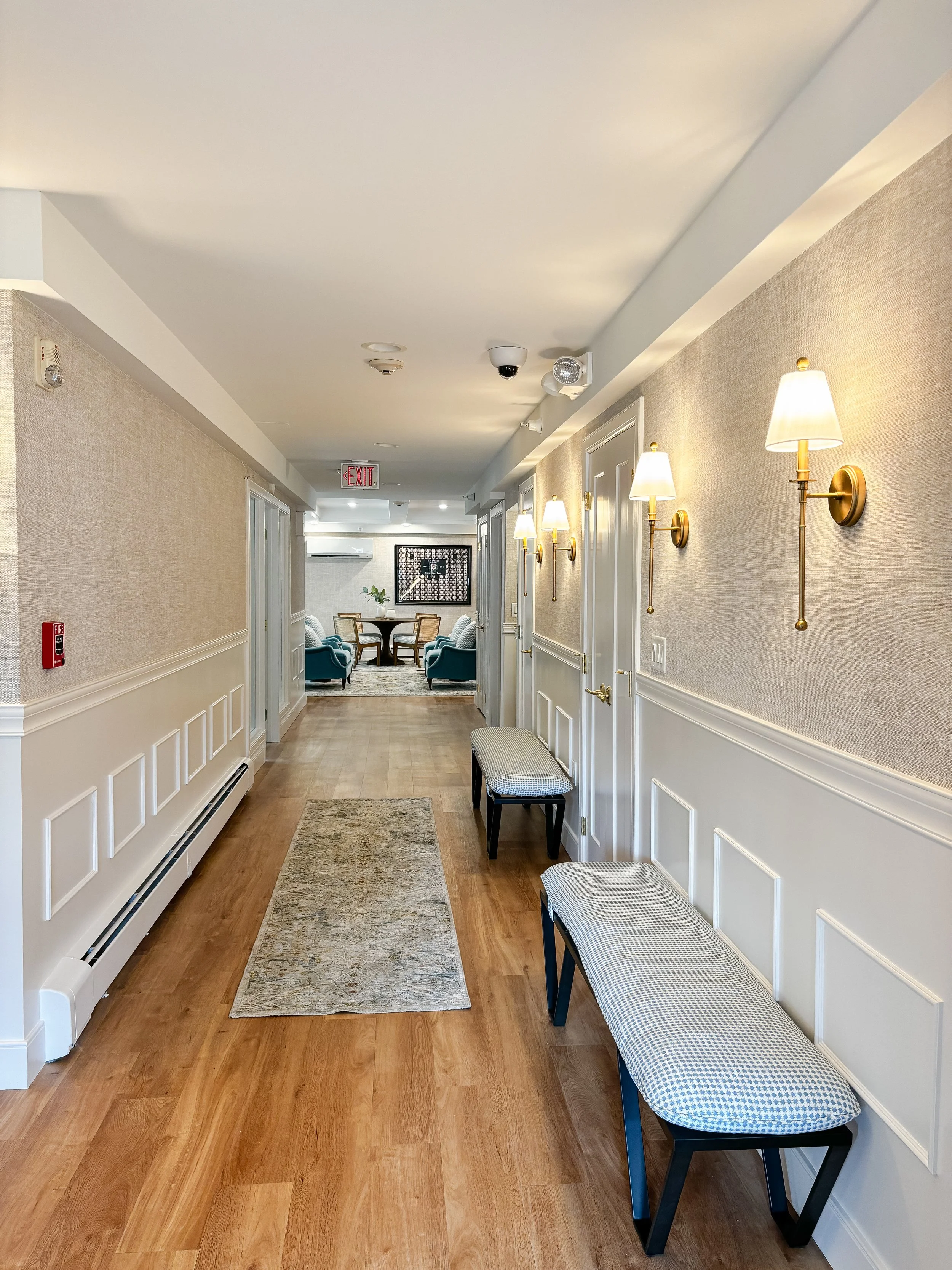 Hallway with wall-mounted lamps, benches, and a view of a lounge area with armchairs and a small table.