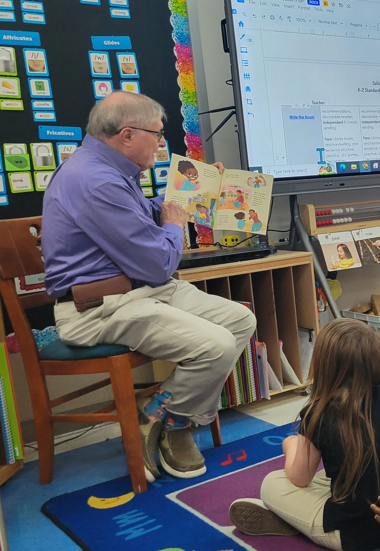 ennis Dulniak of Nana’s Books Foundation reads a children’s book to first-grade students seated on a classroom rug.
