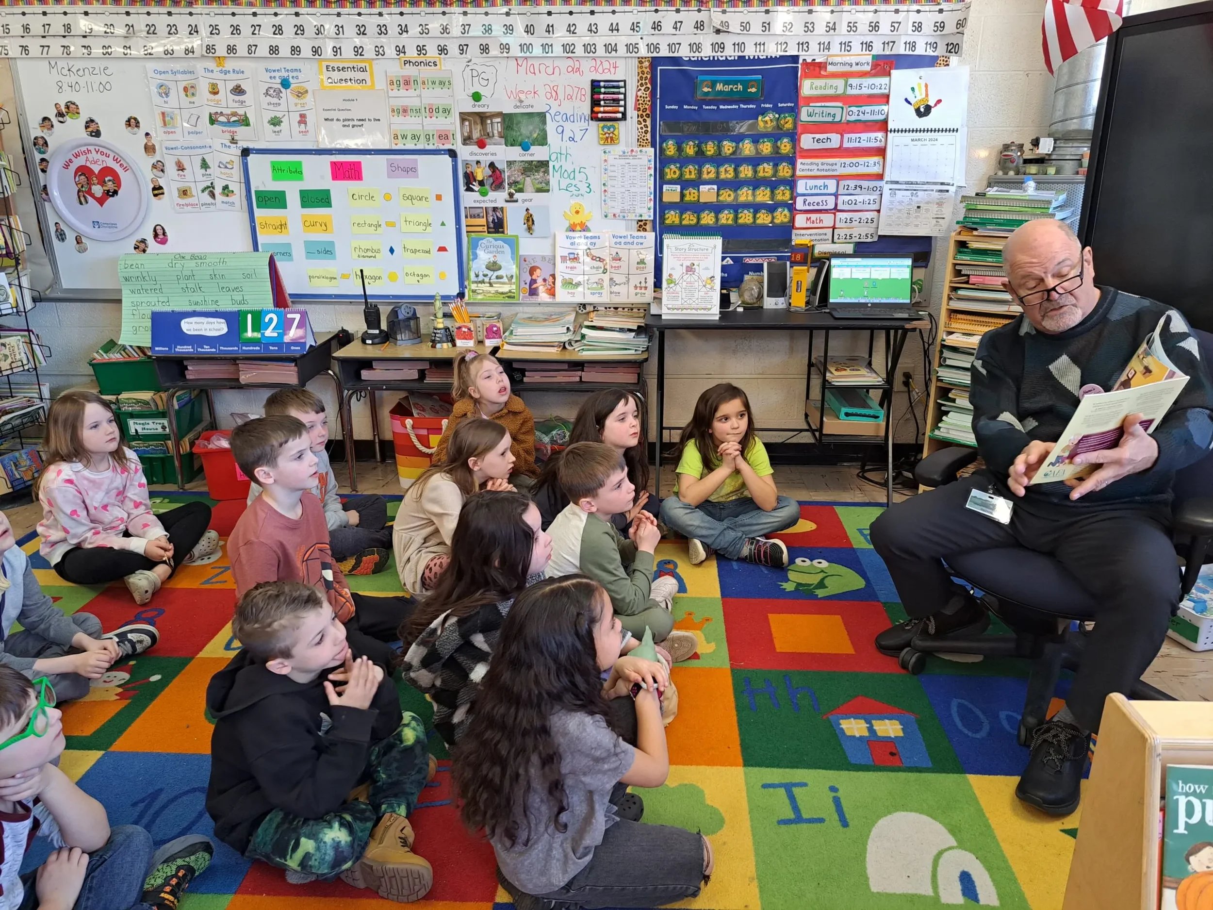 Students gather on a classroom rug as a guest reads a book provided by Nana’s Books Foundation.