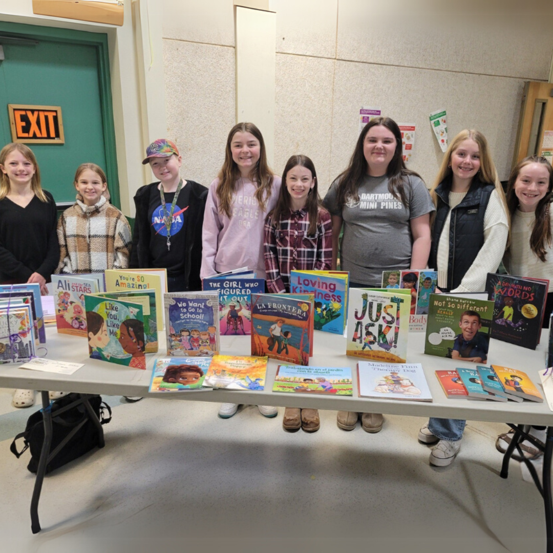 Students stand behind a table displaying children’s books about disabilities, differences, kindness, and inclusion.