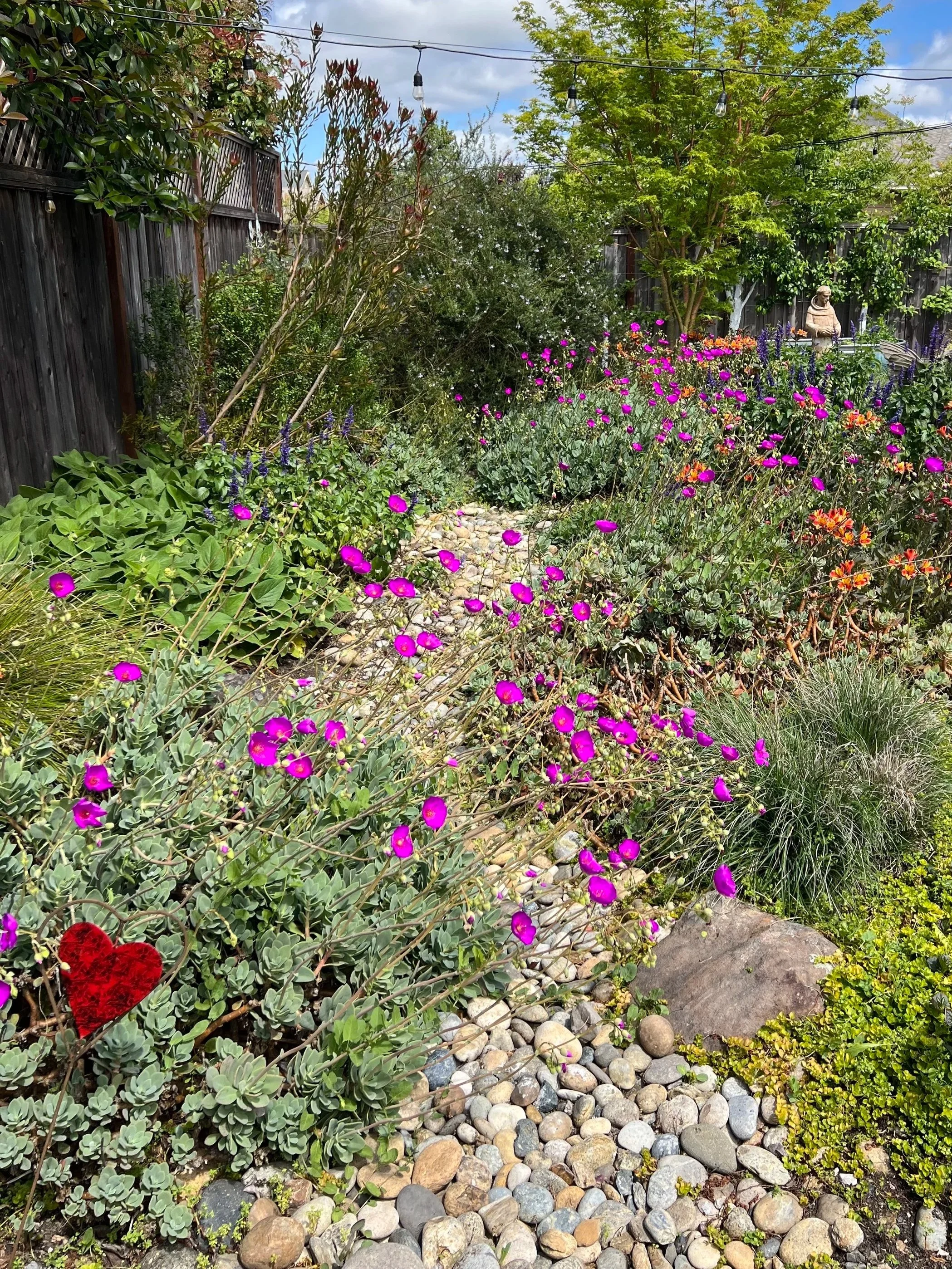 A lush garden with vibrant purple, red, and orange flowers, surrounded by green foliage, a wooden fence, and decorative string lights overhead. A statue of a pig is visible on the right side, standing near a metal bench.