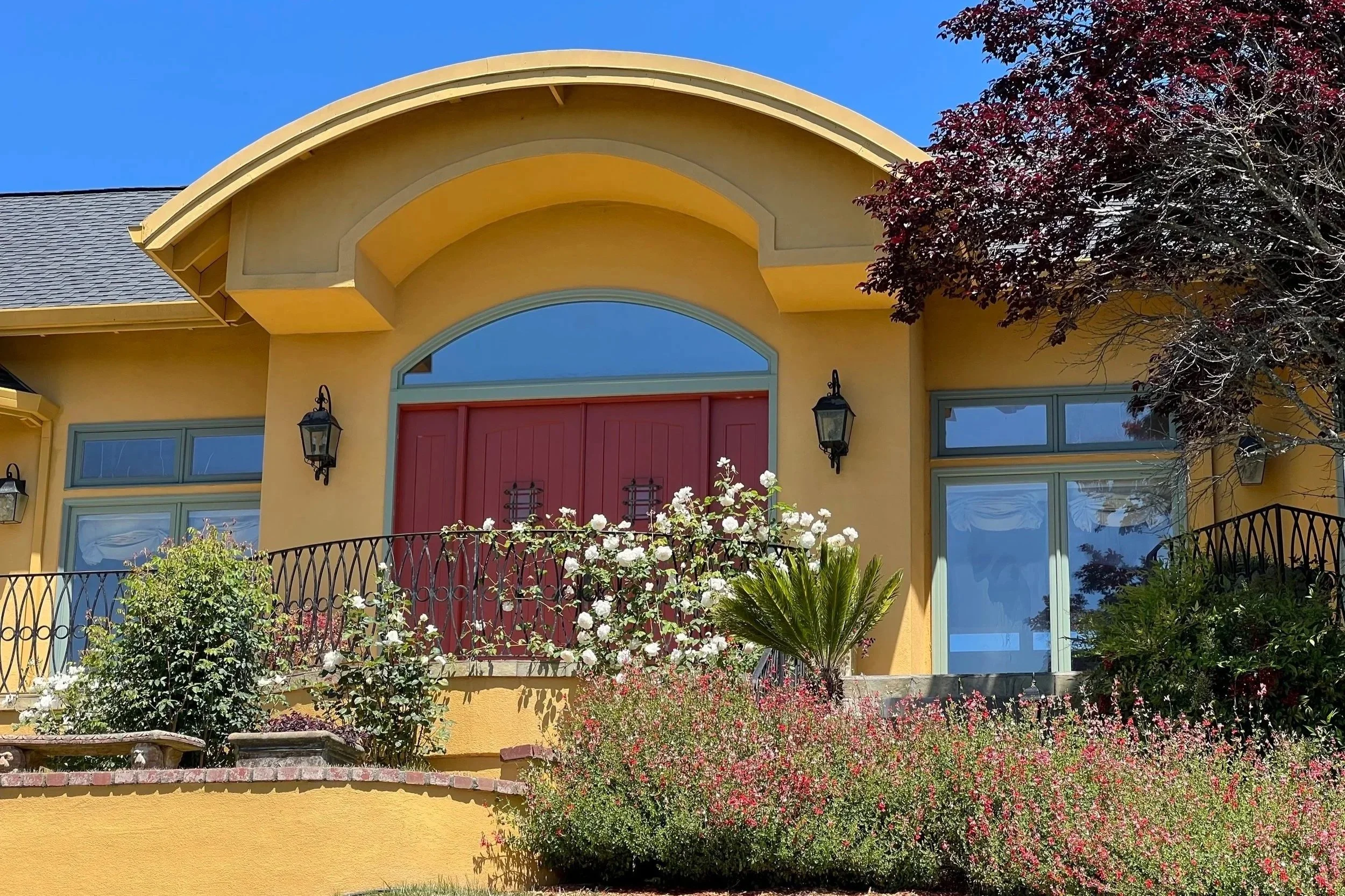 Front view of a yellow house with a red door surrounded by a garden and flowering bushes, blue sky in the background.
