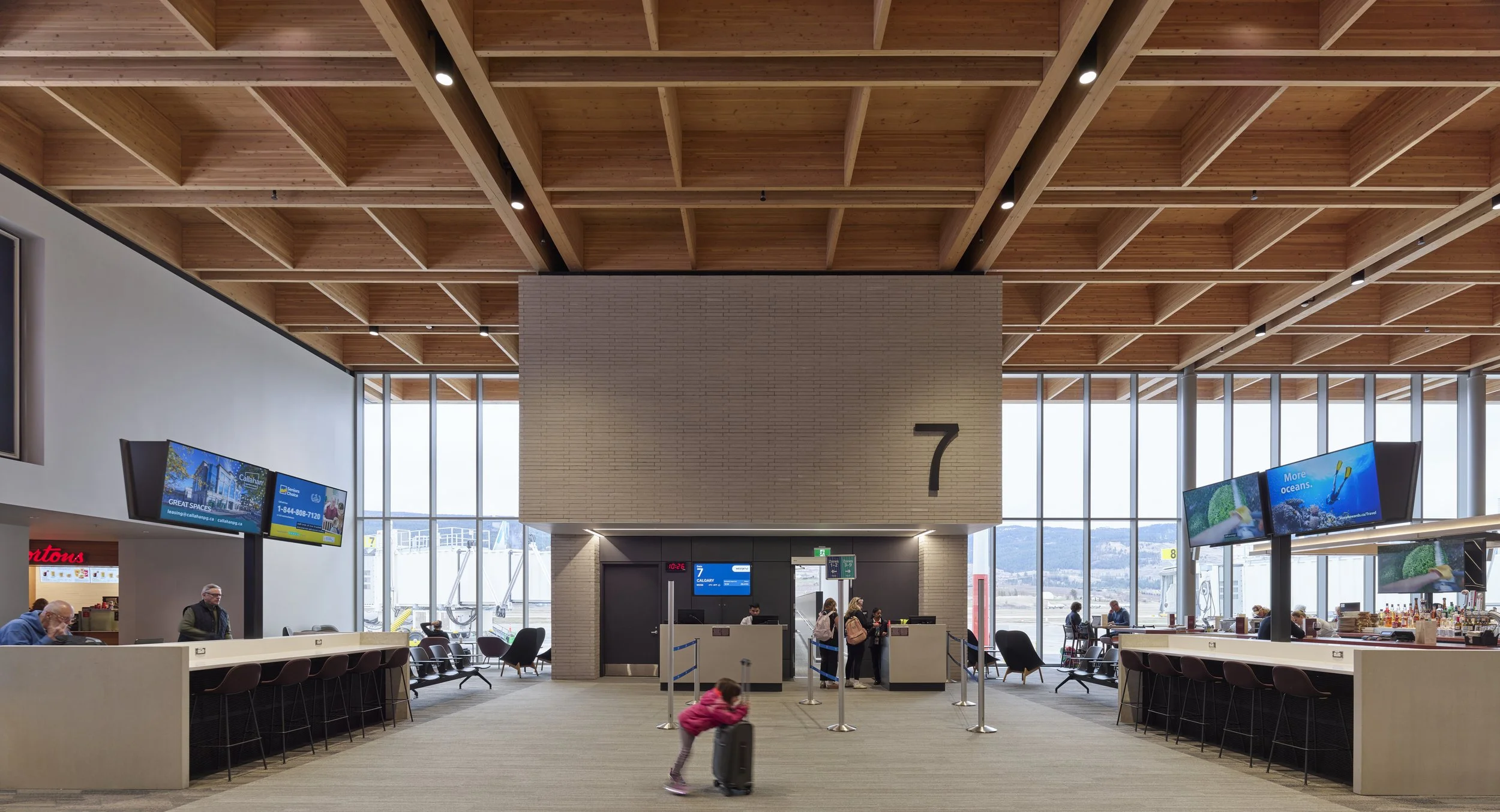 Interior of an airport waiting area with check-in counters and seating. A large window shows airplanes outside. The ceiling has wooden beams, and there are digital screens and a clock visible.