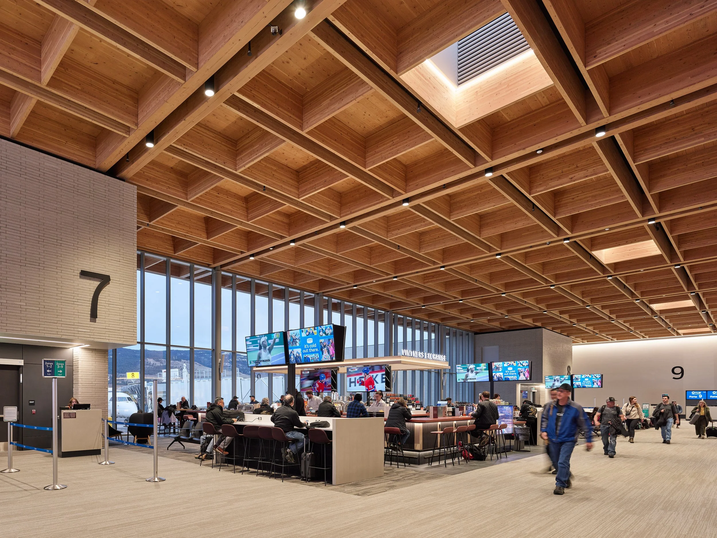 Interior of an airport terminal with high wooden ceiling, digital screens displaying sports news, people waiting and walking around, and large glass windows showing outside view.