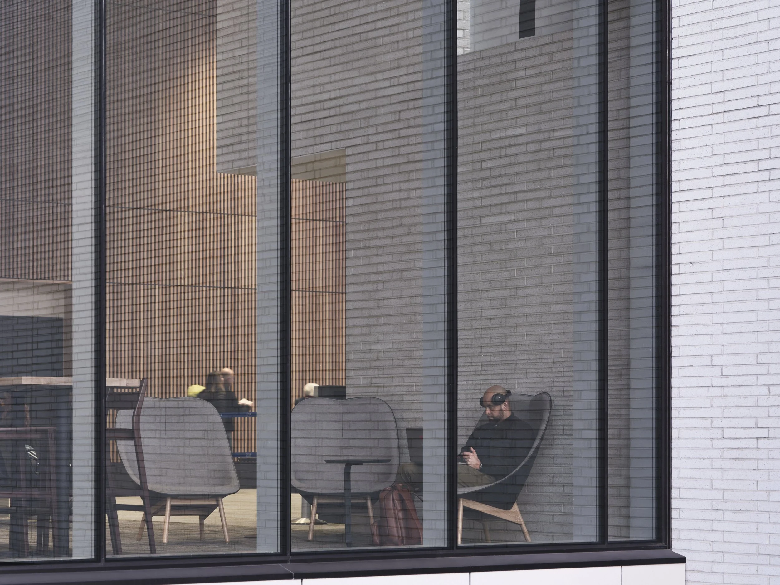 Reflection of a person holding a phone, sitting at a table with chairs outside, seen through a glass building facade.
