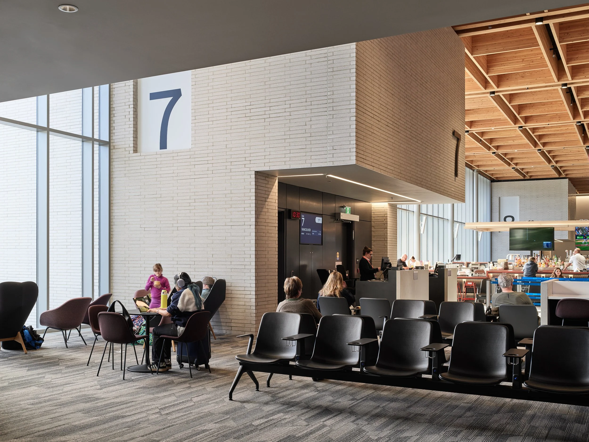 An airport terminal interior with large windows, seating, and a large number 7 sign indicating gate 7. People are sitting and waiting, some with laptops or phones. A person is walking with a yellow water bottle, and a few airline staff are working ne