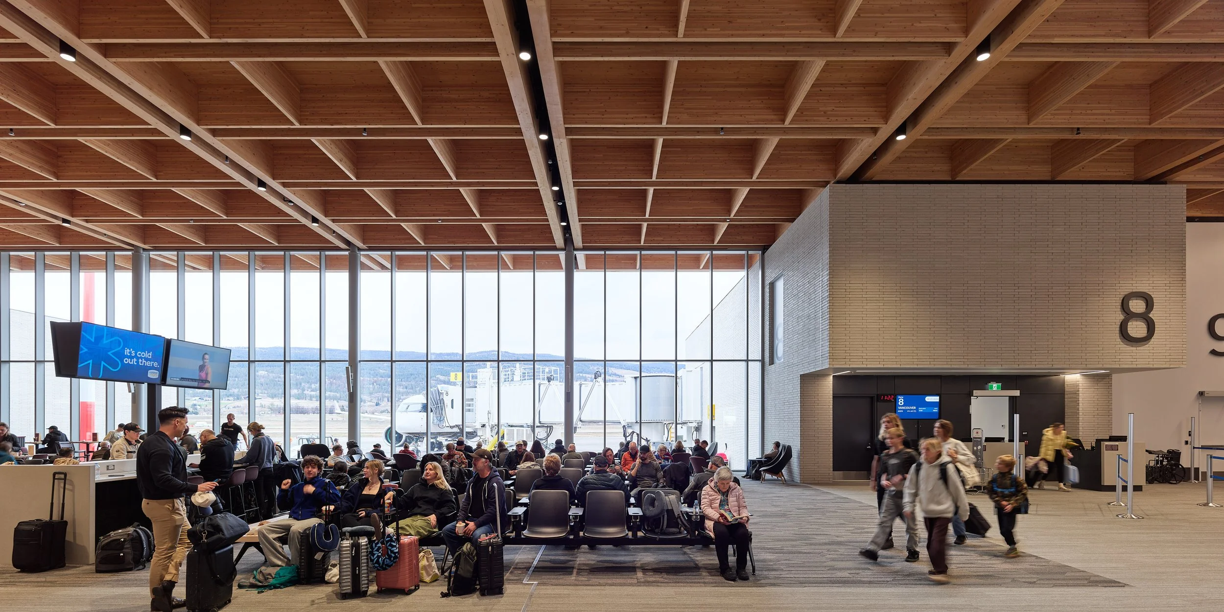Passengers sitting and walking at an airport gate with large windows and some luggage, waiting for flights.