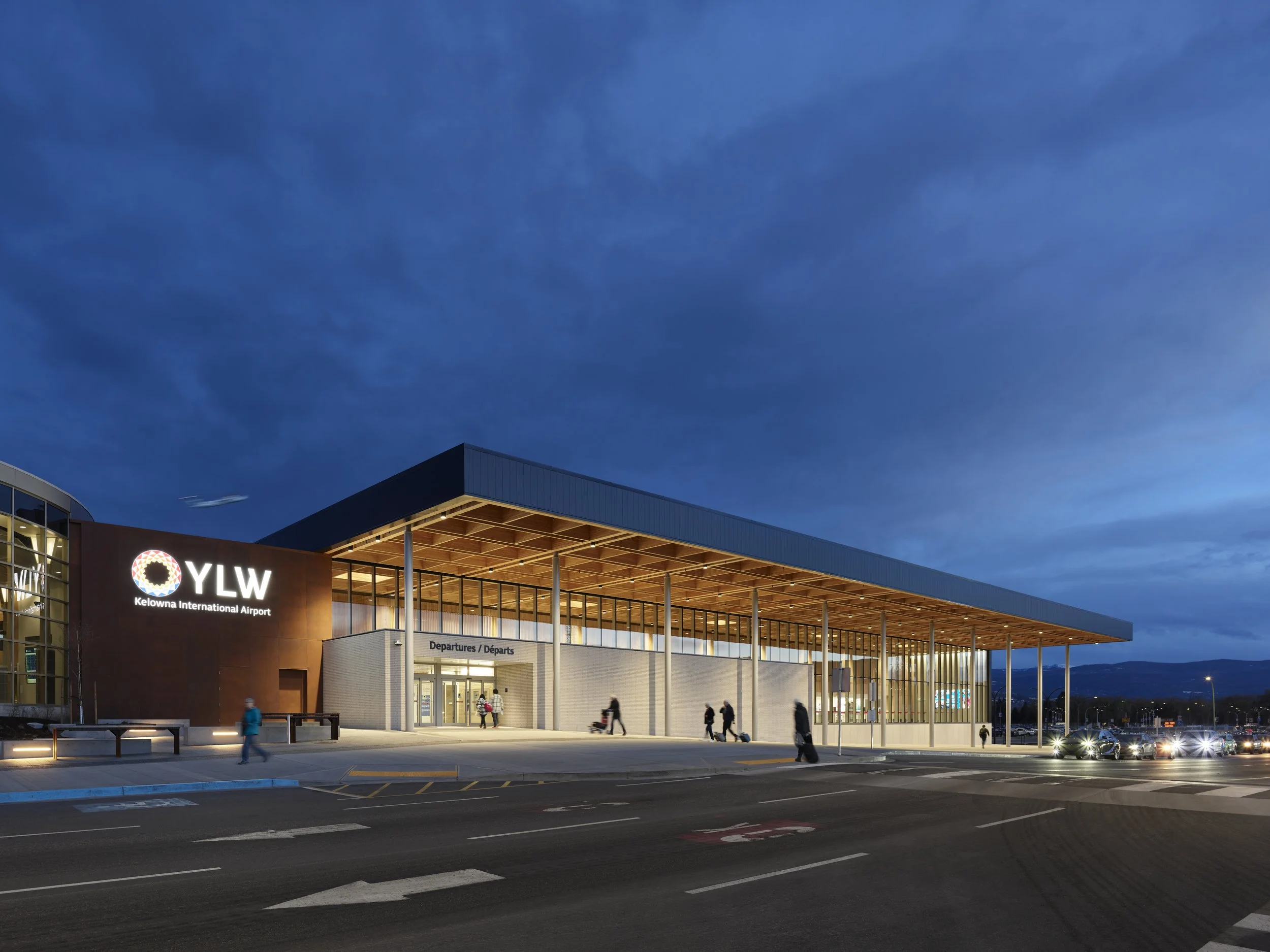 Nighttime exterior view of Kelowna International Airport terminal with people walking outside, lit building, and dark sky.