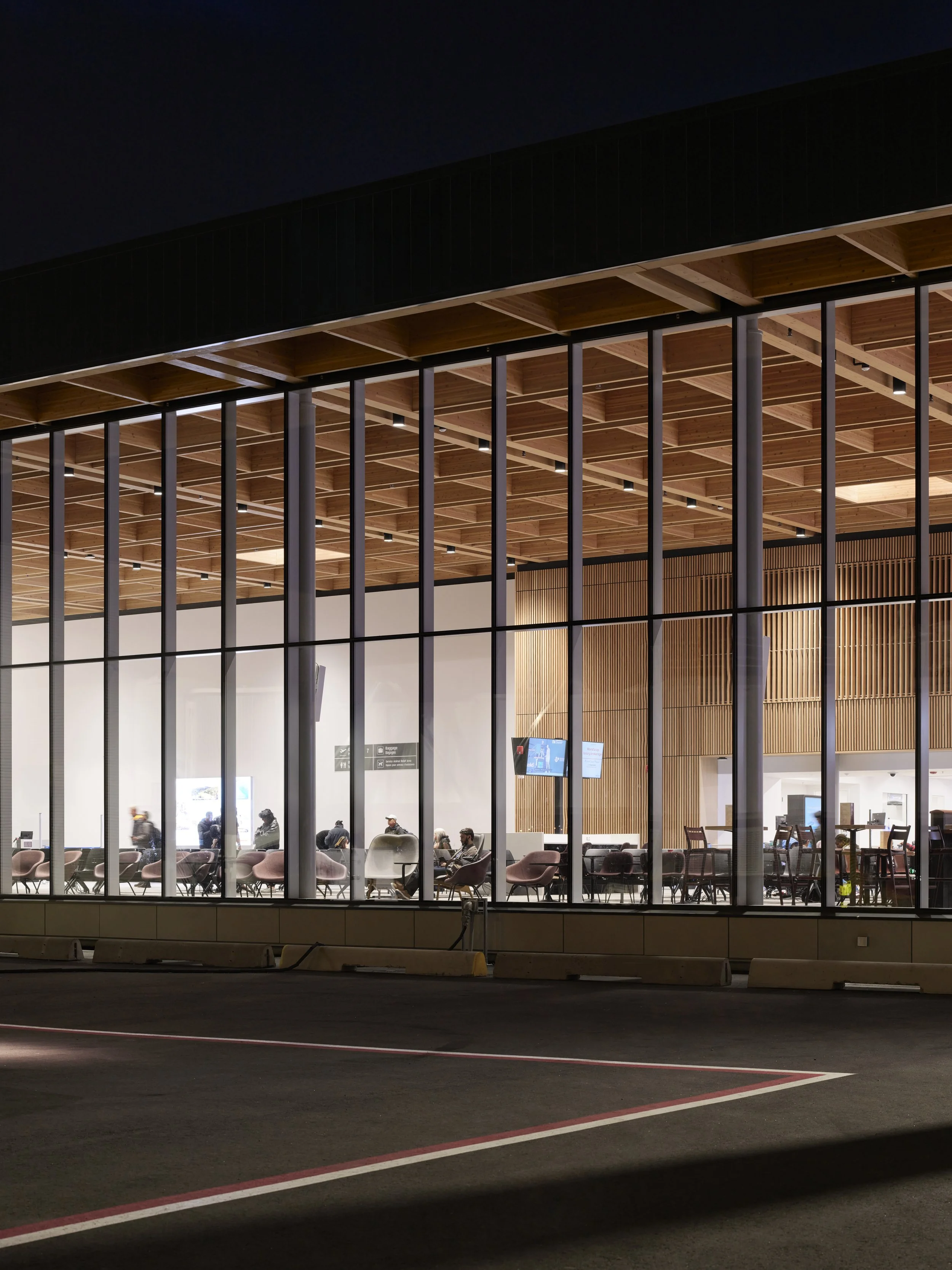 Night view of an airport terminal with large glass windows showing the interior, including waiting seats and people, and a modern wooden ceiling.