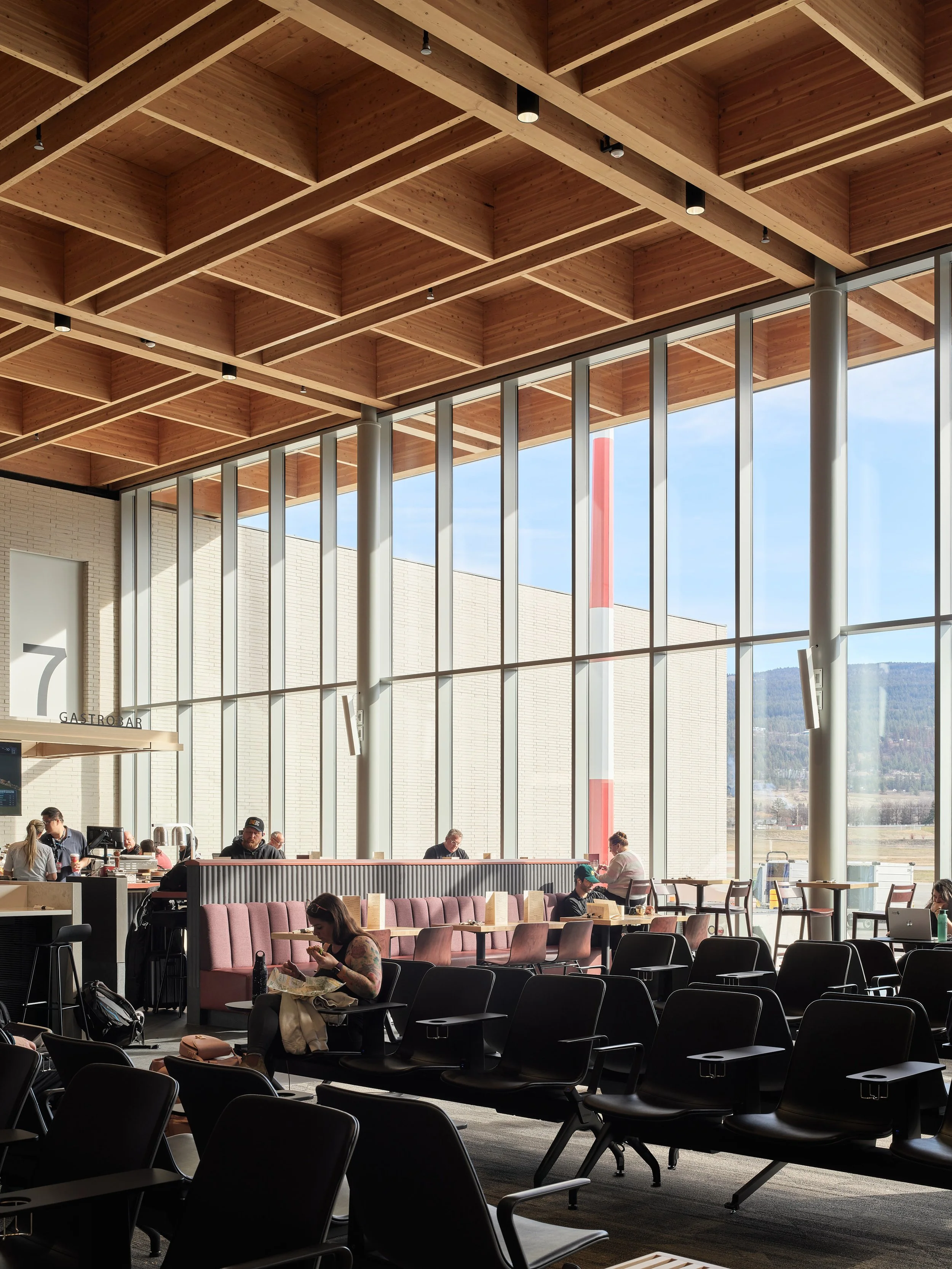 Interior of an airport lounge with large windows showing an outdoor landscape, several people sitting and waiting, some using laptops or phones, with a ceiling featuring wooden beams and modern lighting.