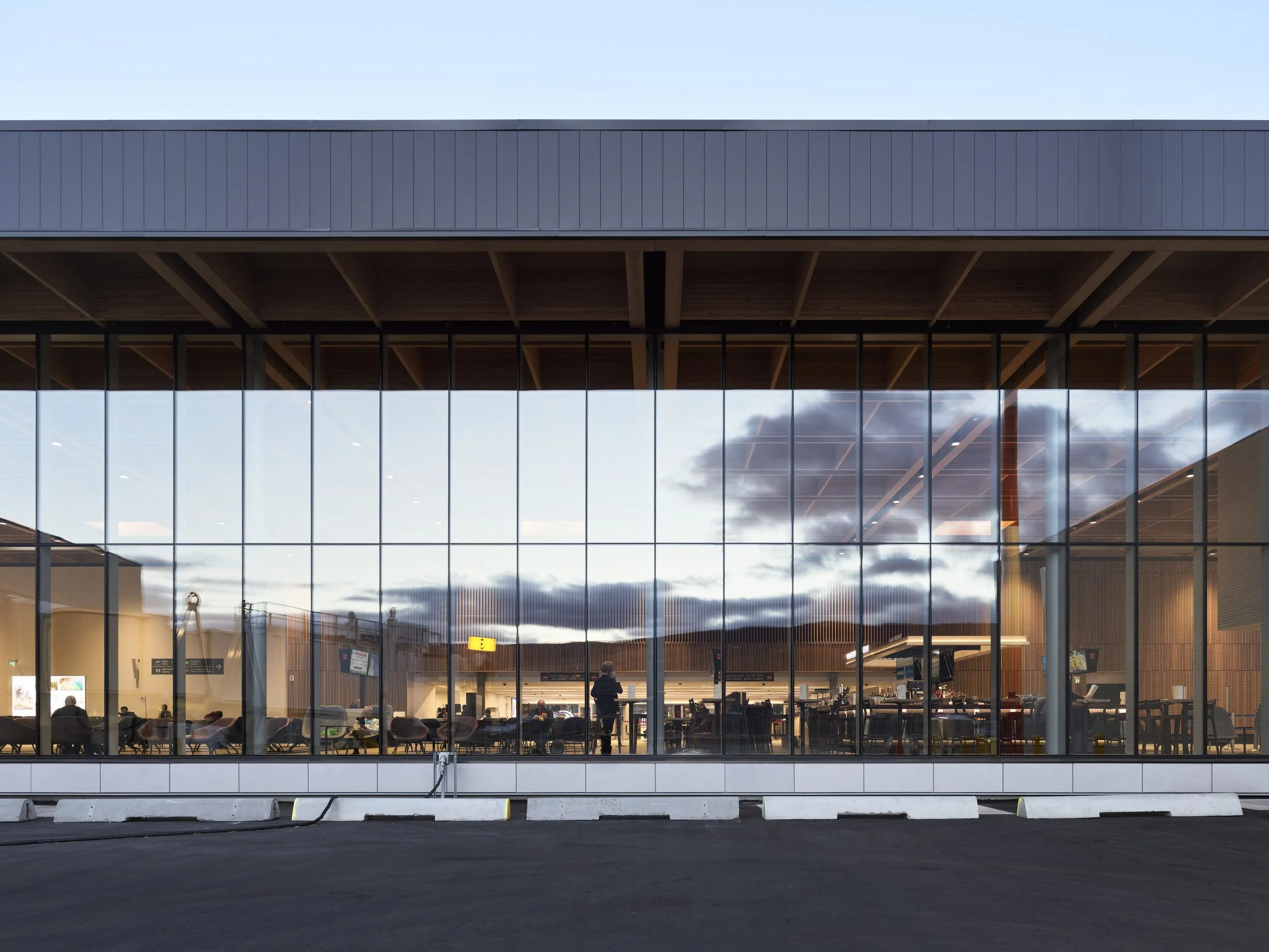 View of an airport terminal with large glass windows showing the interior where multiple passengers are waiting, some sitting and some standing, and the ceiling inside has wooden beams. The exterior has a clear sky with scattered clouds.