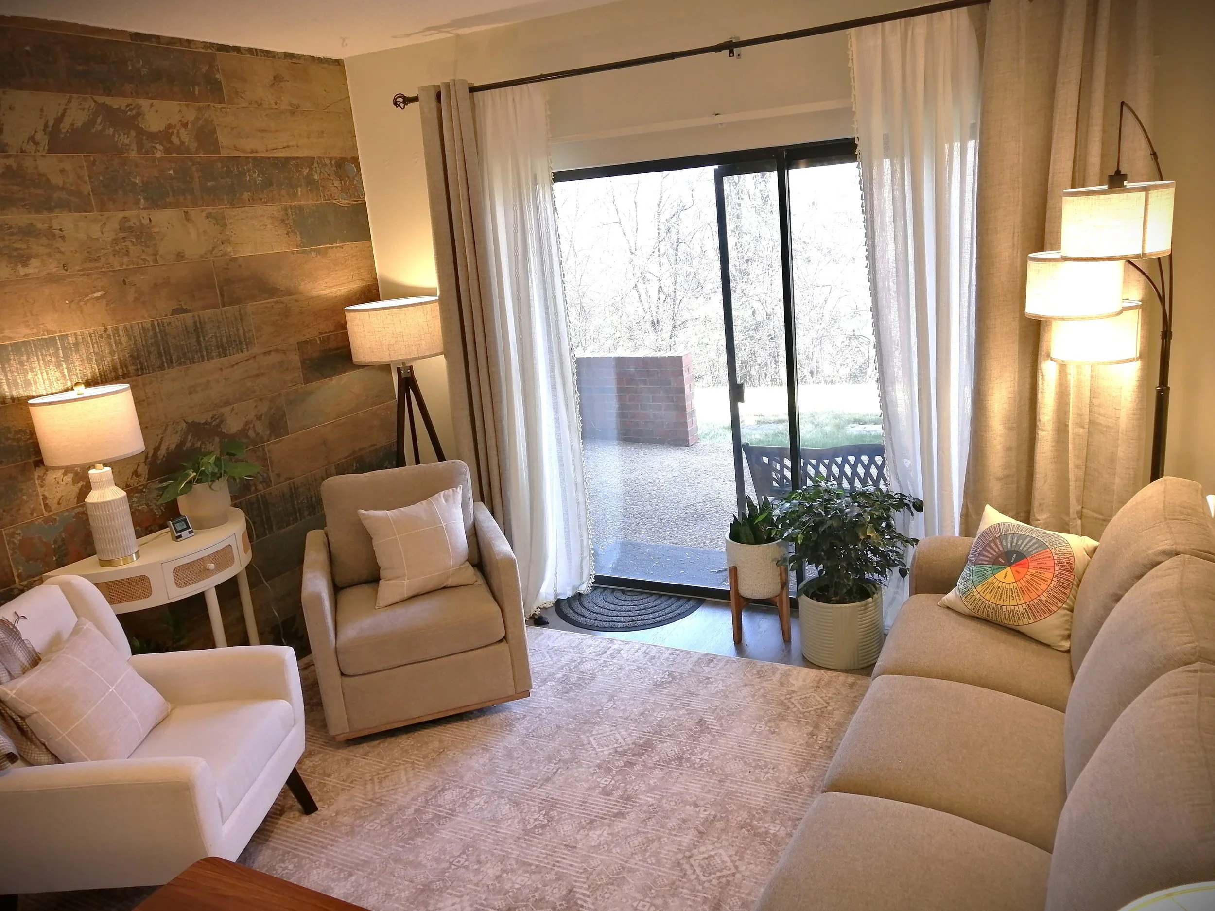 Living room with beige sofa, armchair, side table, and potted plants near sliding glass door with white curtains, wood accent wall, and floor lamps.