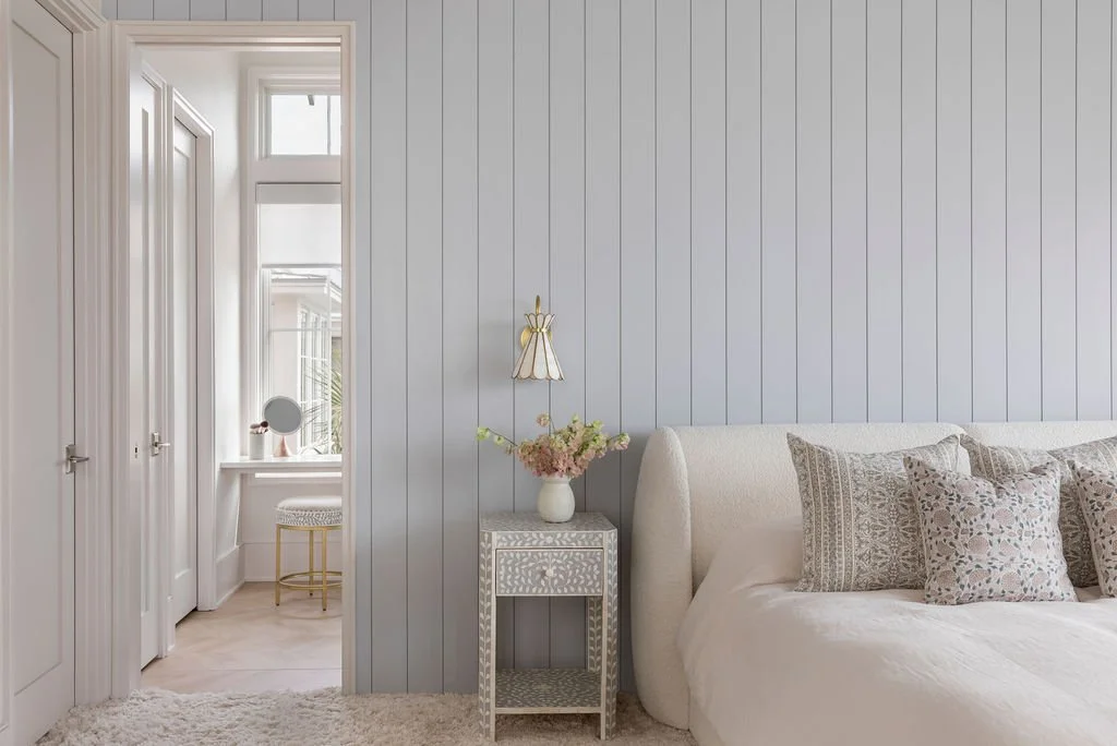 A bedroom with a bed, decorative pillows, a nightstand with a vase of flowers, and a wall-mounted lamp by Charleston interior stylist Ellen Casey