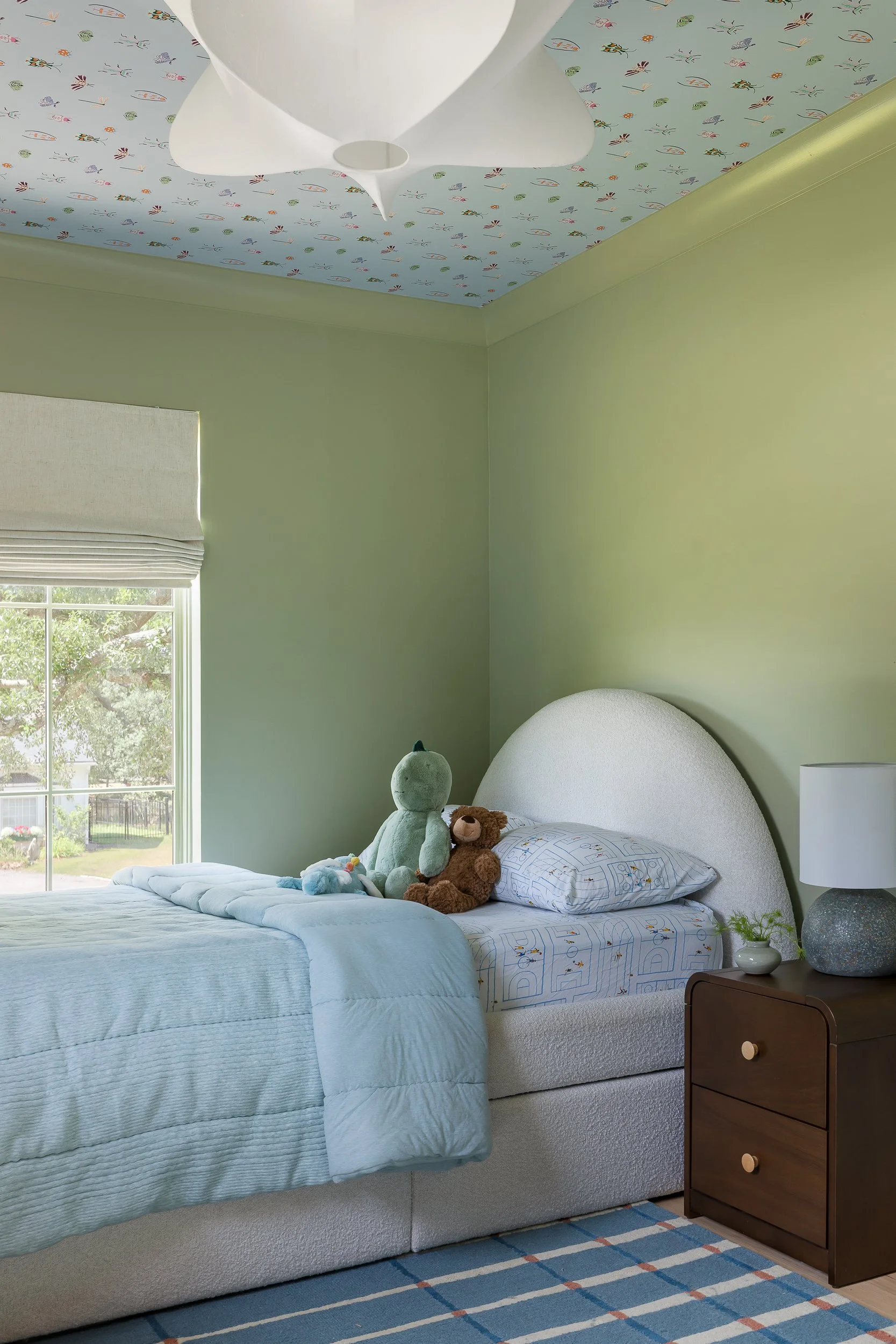 A bedroom with a green wall, a window with a white Roman shade, a bed with a blue quilt, stuffed animals, and toys. There is a brown nightstand with a lamp and a small potted plant. The ceiling has light blue wallpaper with a whale, fish, and crab pa