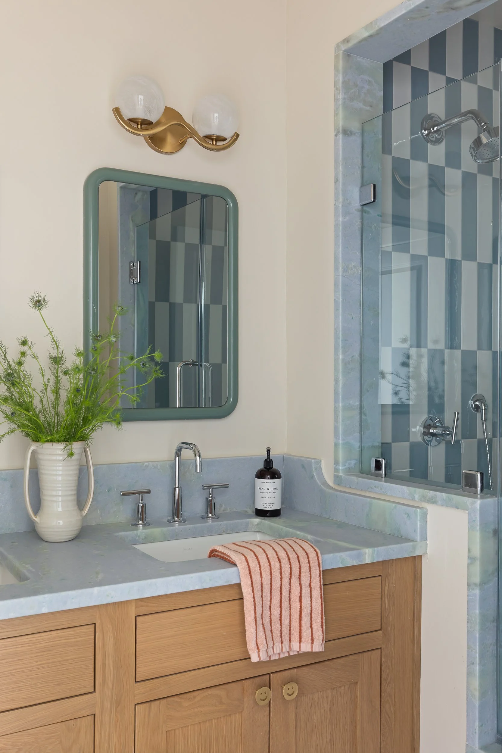 Bathroom vanity with a light wood cabinet, marble countertop, white sink, and silver fixtures. A striped pink towel hangs over the front of the cabinet. A potted greenery on the counter, with a mirror and a light fixture above. A soap bottle and patt