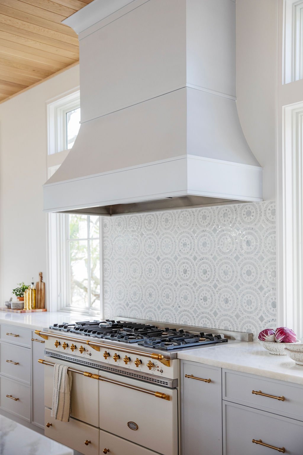 A modern kitchen with a white stove and oven, white cabinets with gold handles, a patterned backsplash, and a white range hood.