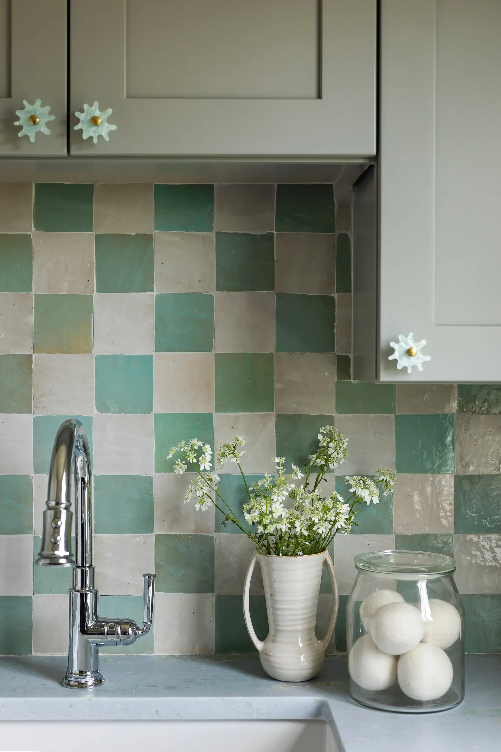 A kitchen countertop with a stainless steel sink and a silver faucet. Behind the sink, there are green and beige checkered tiles on the wall. A white vase with green foliage and small white flowers is placed on the counter, along with a transparent j