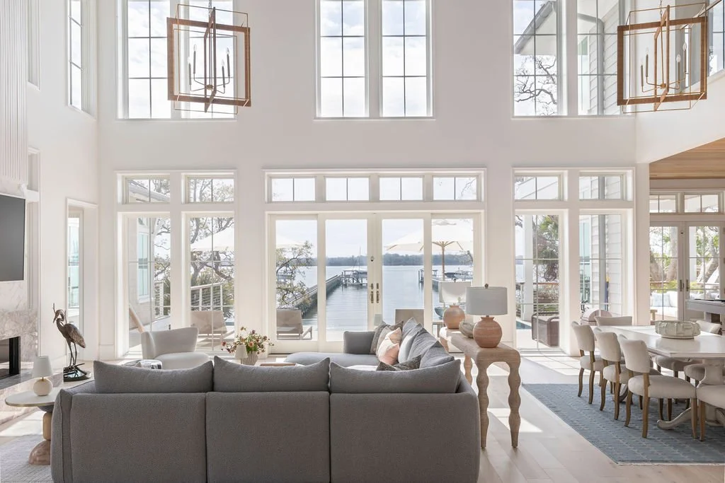 Bright living room with large windows and a view of a dock and water outside. Features a gray sofa, white chairs, wooden side tables, and a dining area.