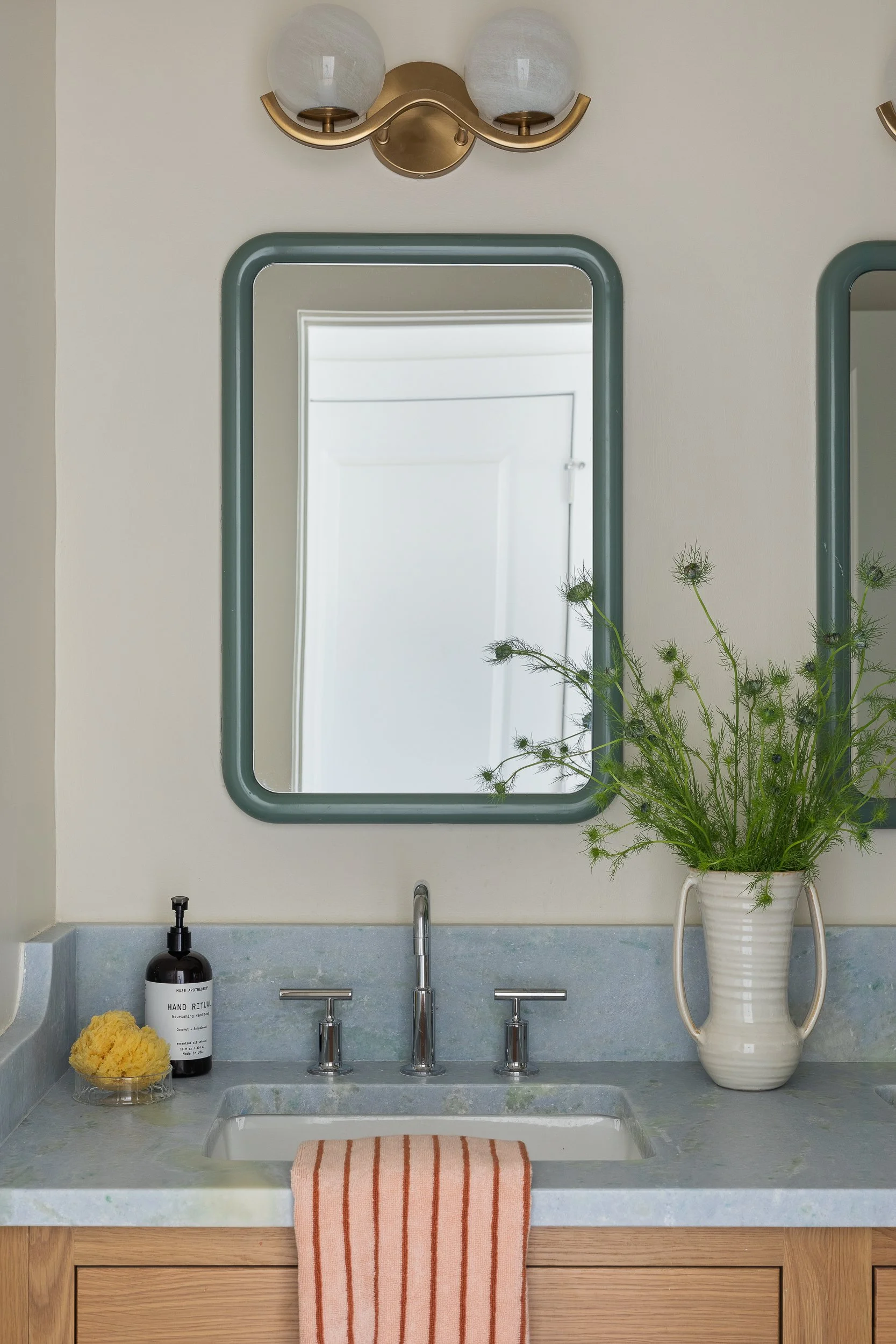 Bathroom sink with a small mirror, a potted plant, a soap dispenser, moisturizer, and yellow sponge, with a striped towel hanging over the sink, wooden cabinet below, and a light fixture with two white globes above.
