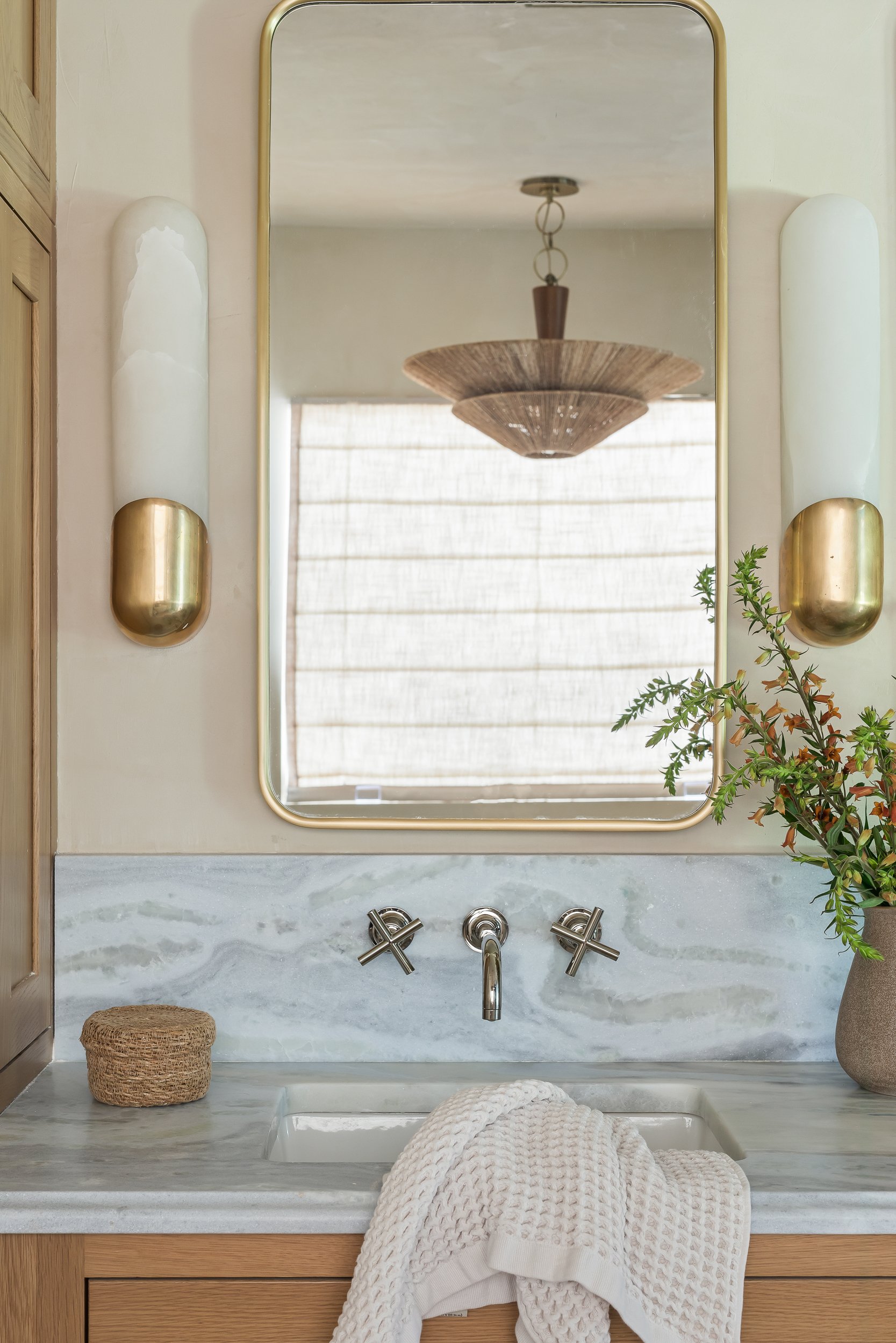A bathroom sink with a white textured towel draped over the edge, a woven basket on the left, and a gray stone countertop; a mirror with a gold frame above the sink, reflecting a wooden chandelier and a window with blinds; wall sconces on either side