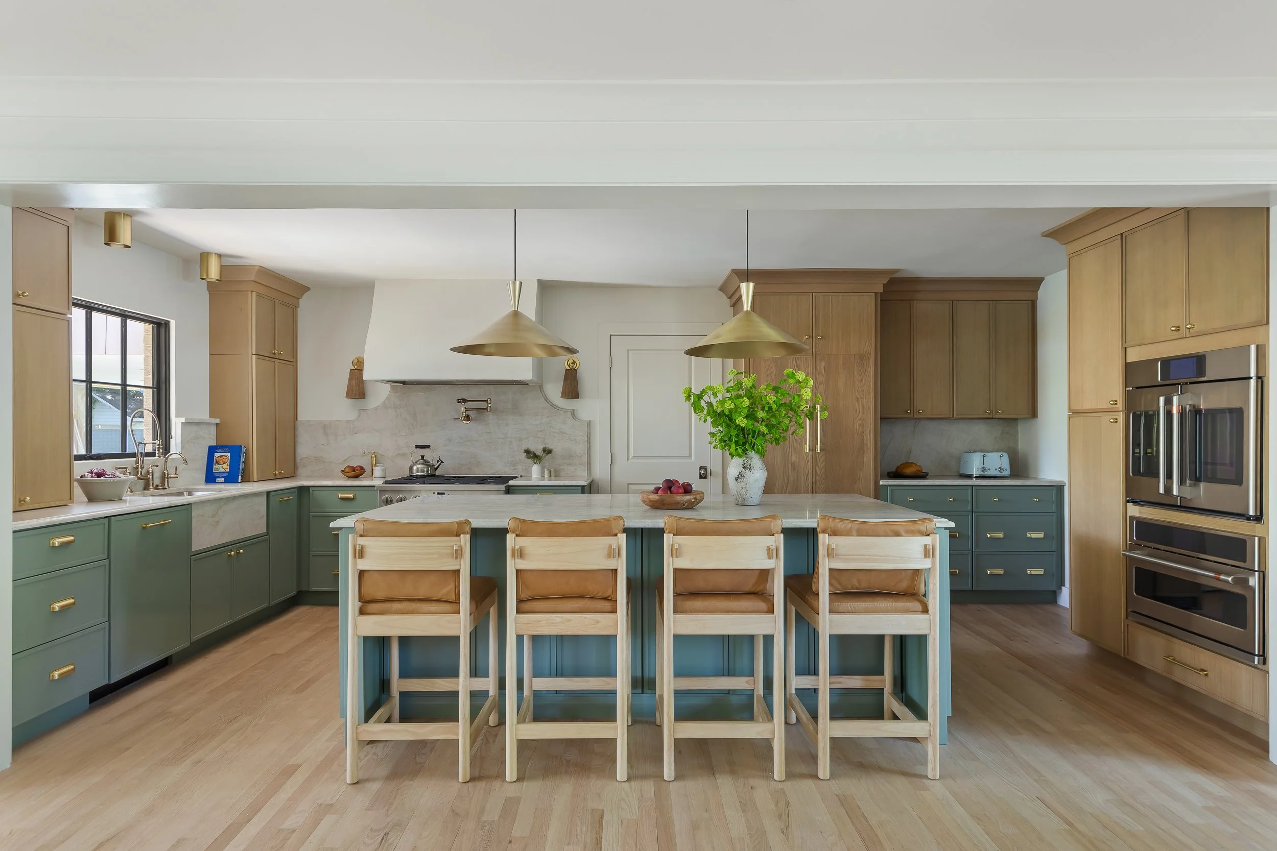 Modern kitchen with green and wood cabinetry, white marble countertops, and a large island with a vase of green plants and red apples, featuring brass pendant lights.