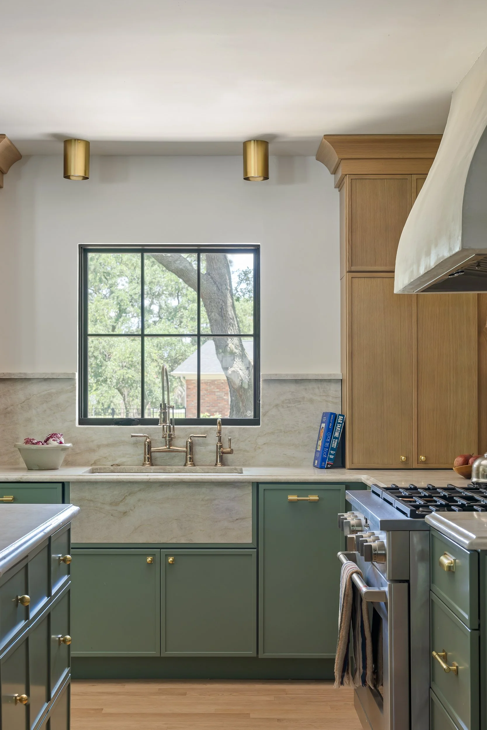 A modern kitchen with green cabinets, marble countertops, a window with a view of trees outside, and brass fixtures. There are gold cylinder ceiling lights and a wooden cabinet on the right.