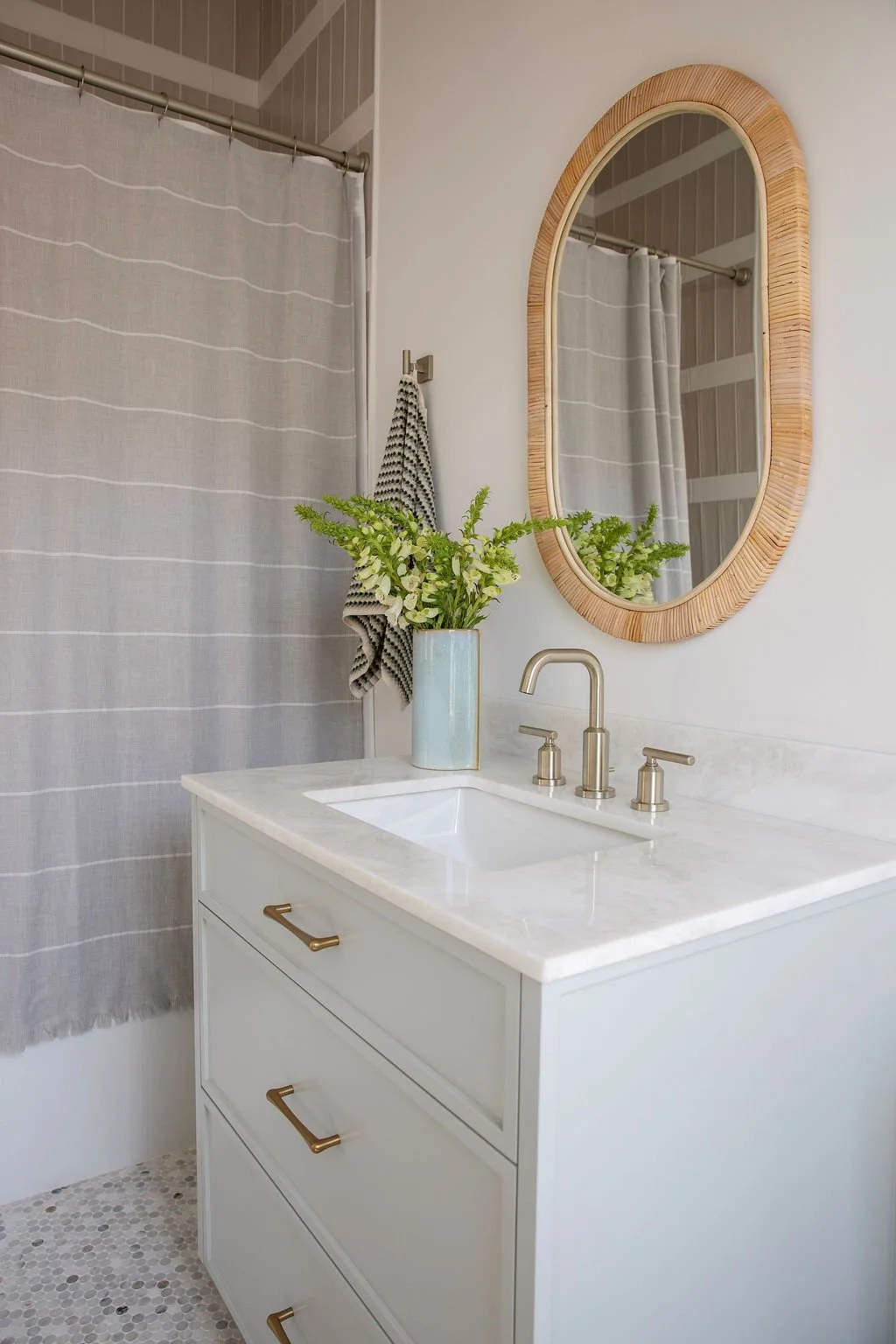 A bathroom vanity with a white countertop, a sink, and three drawers with brass handles. A tall vase with green foliage and white flowers is on the counter. A round wooden mirror is mounted on the wall above the sink, and a striped towel hangs on a h