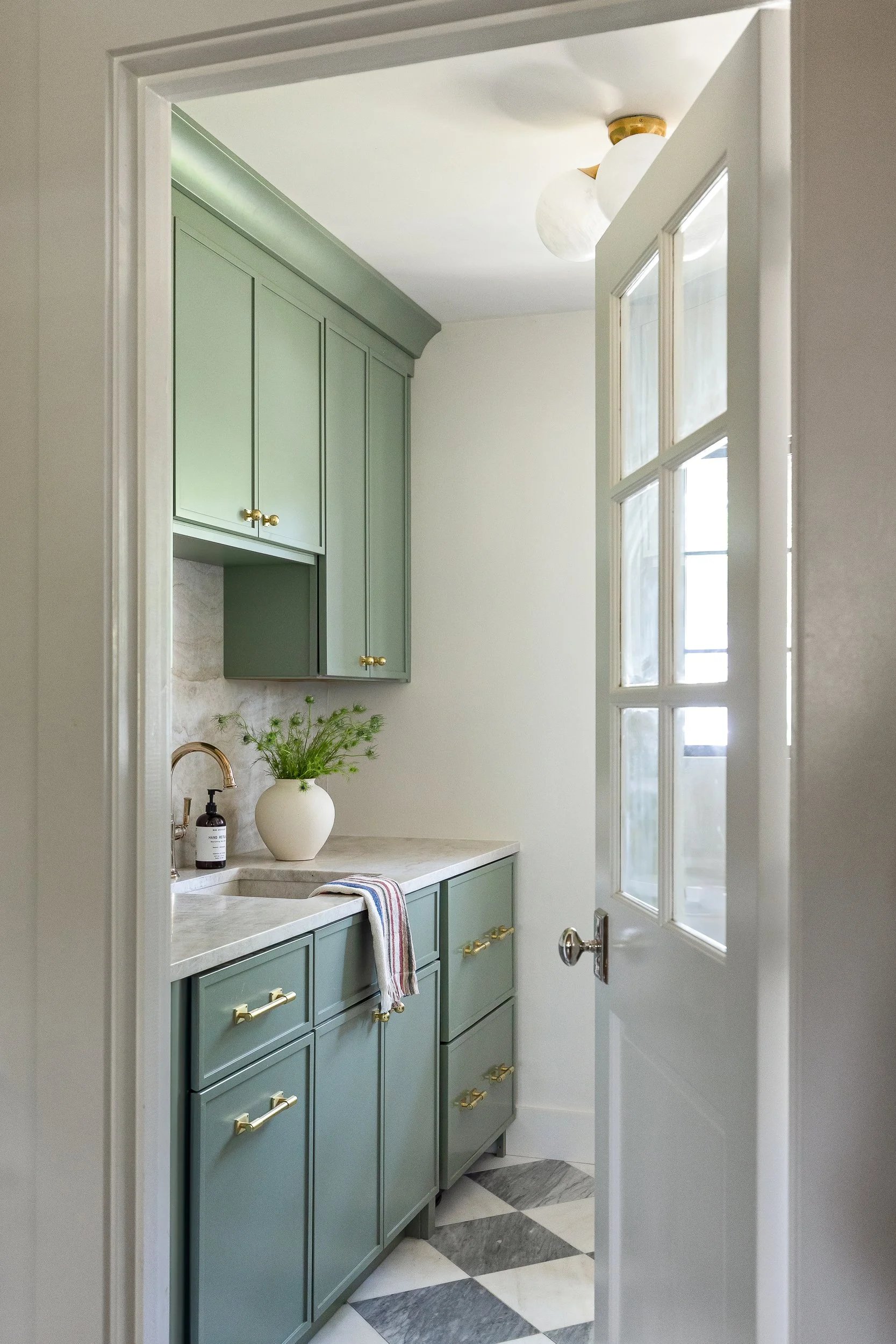 A small bathroom with light green cabinets, a beige countertop, a white vase with green plants, a striped towel, a soap dispenser, and a window with multiple panes in a white frame. The floor has black, white, and grey marble tiles, and there's a cei