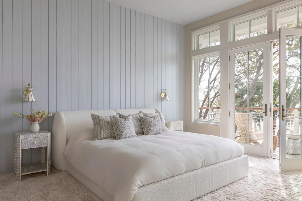Bright bedroom with light blue shiplap accent wall, white upholstered bed with decorative pillows, side tables, and a balcony with outdoor seating visible through large glass doors and windows.