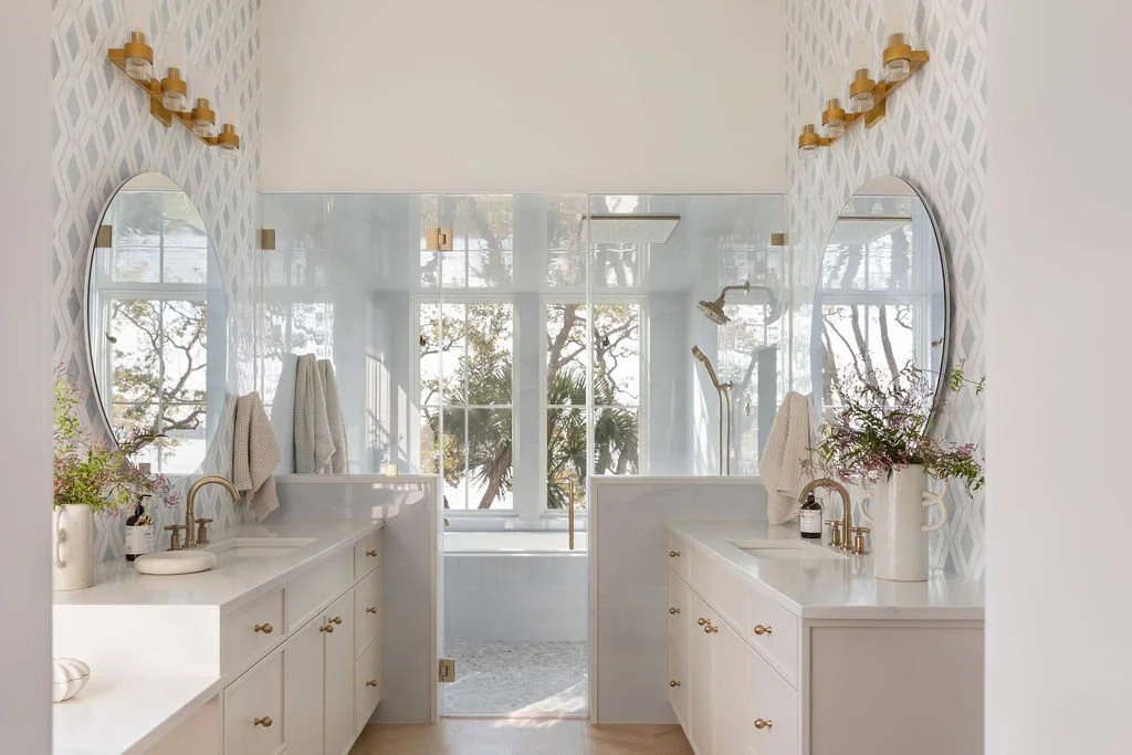 Dual bathroom vanity with white cabinets, gold handles, and oval mirrors. Each sink has a gold faucet, with a vase of flowers on each side. The bathroom has a large window, natural light, and a white patterned wall.