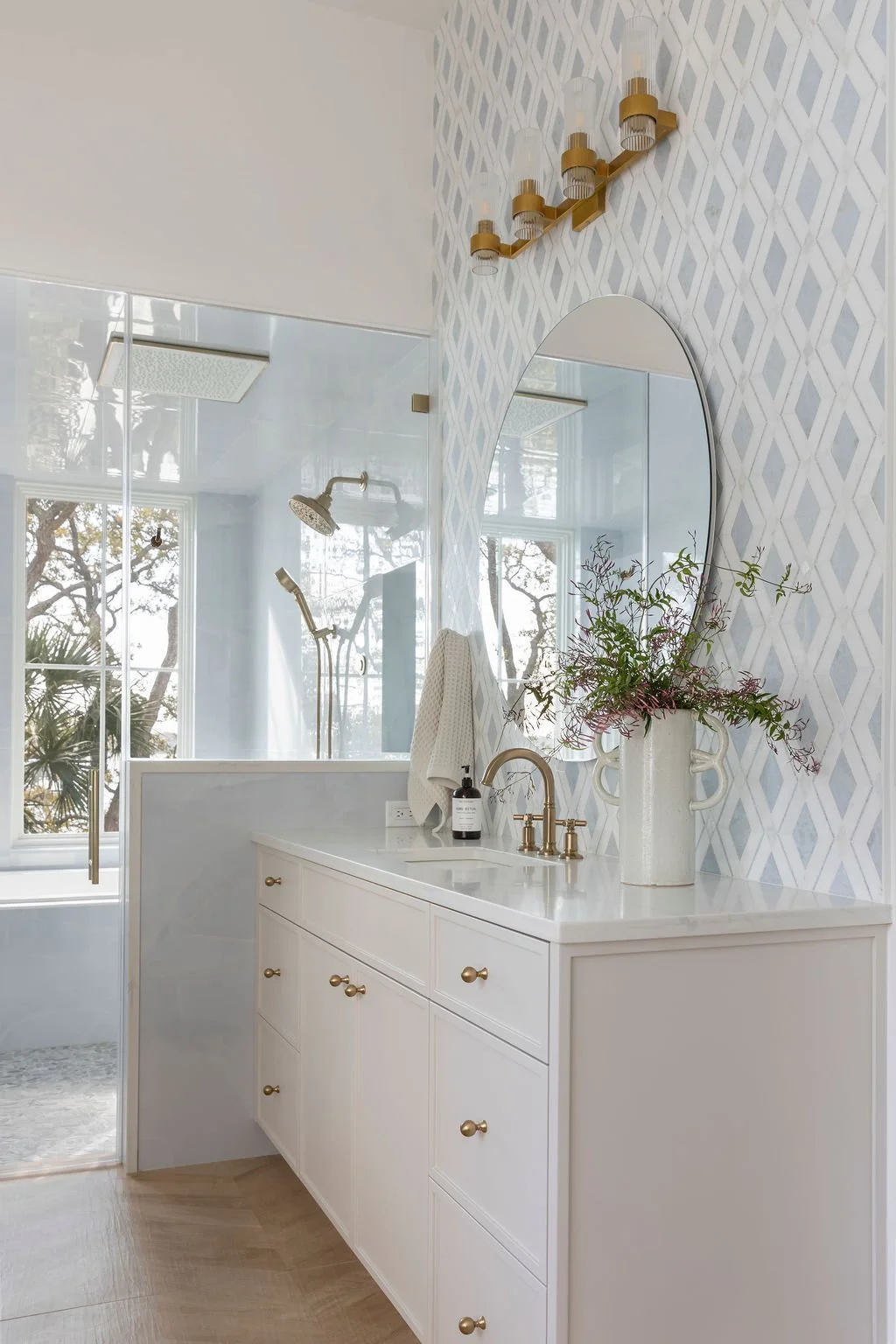 Bathroom vanity with white cabinet, gold hardware, oval mirror, and a vase with greenery on the countertop; patterned wallpaper on the wall, reflection of a shower with glass door, window with trees outside, and gold light fixture with multiple bulbs