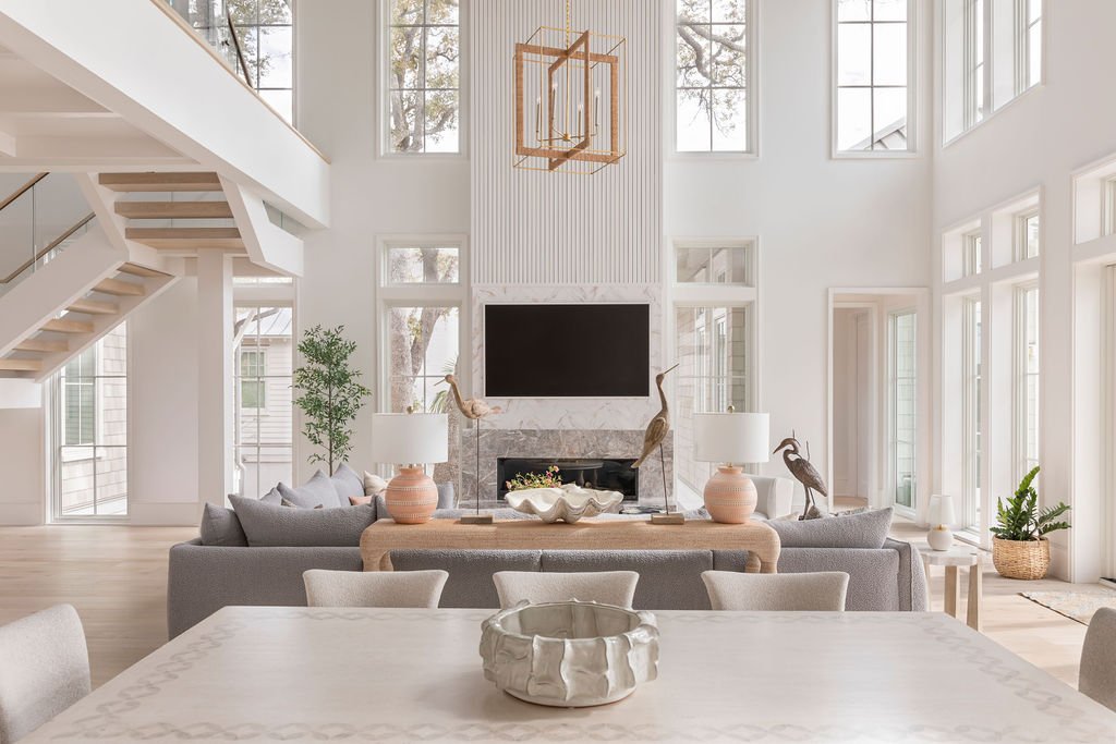 Bright, modern living room with large windows, a white fireplace, a flat-screen TV above the fireplace, gray sectional sofa, decorative lamps, potted plants, and nature-inspired decor.