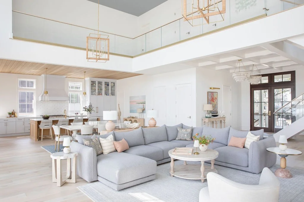 Bright open-concept living room and kitchen with white walls, wood flooring, light gray sectional sofa, decorative pillows, round coffee table with flowers, modern side tables with lamps, and kitchen area with white cabinets and large windows.