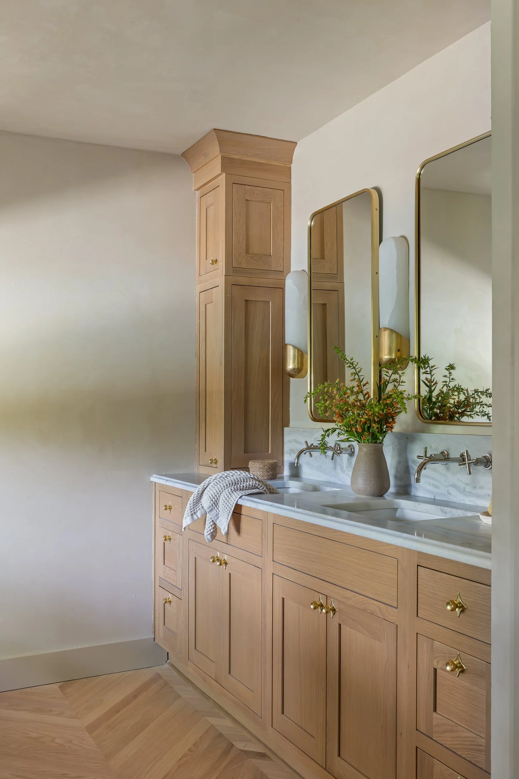 A bathroom vanity with wooden cabinets, a marble countertop, a sink, a potted plant, and two mirrors with gold frames. A towel is draped over the counter.