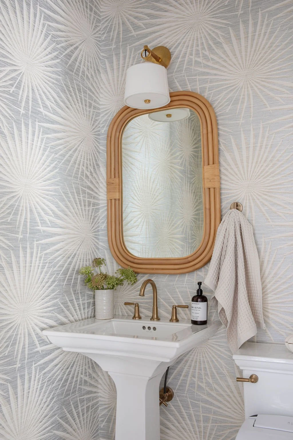 Powder room designed with palm leaf patterned wallpaper, a rectangular mirror with a rattan frame, a white pedestal sink with brass fixtures by Charleston interior stylist Ellen Casey