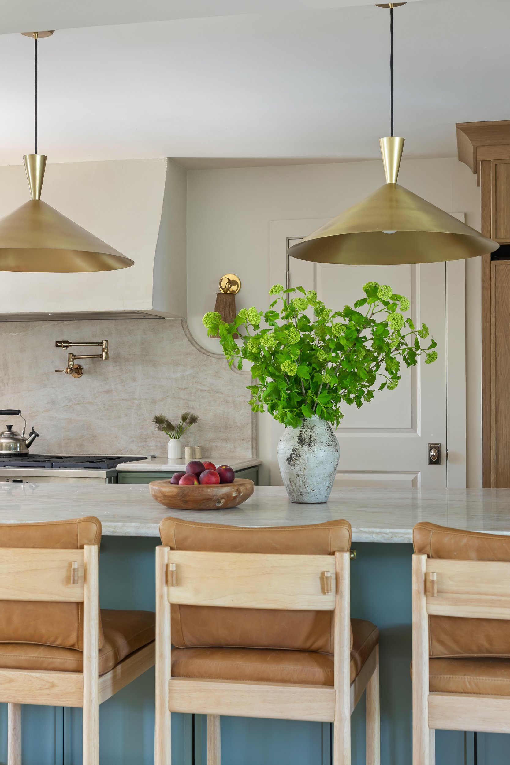 A kitchen interior with a marble countertop, a large potted green plant, wooden chairs, hanging gold pendant lights, and a small wall decoration by Charleston interior stylist Ellen Casey