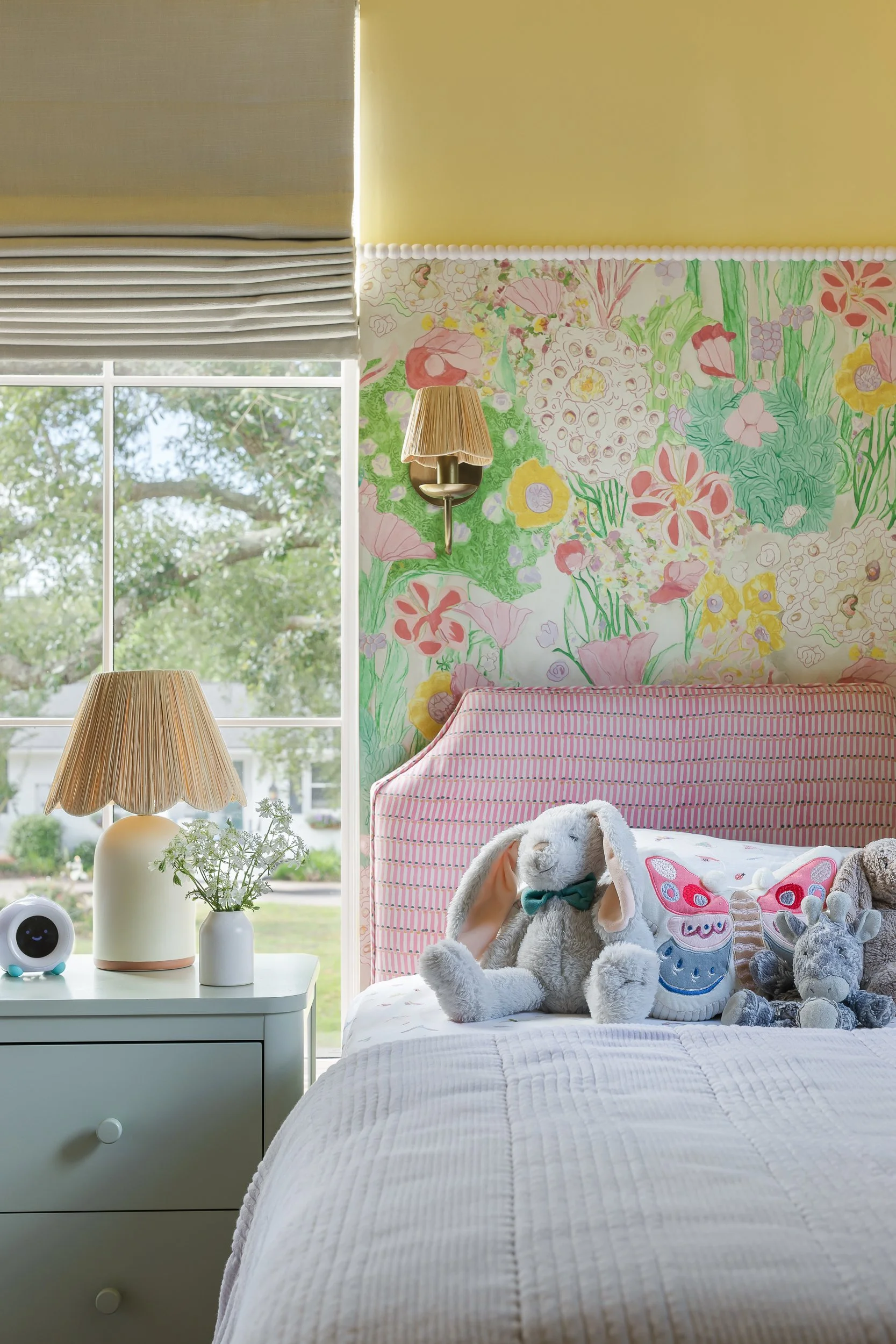 Brightly lit bedroom with large window showing green trees outside. A white nightstand with a lantern-style lamp, small plant, and a round speaker. A soft, stuffed bunny with a teal bow on the bed, along with other plush toys, and a pink-striped head